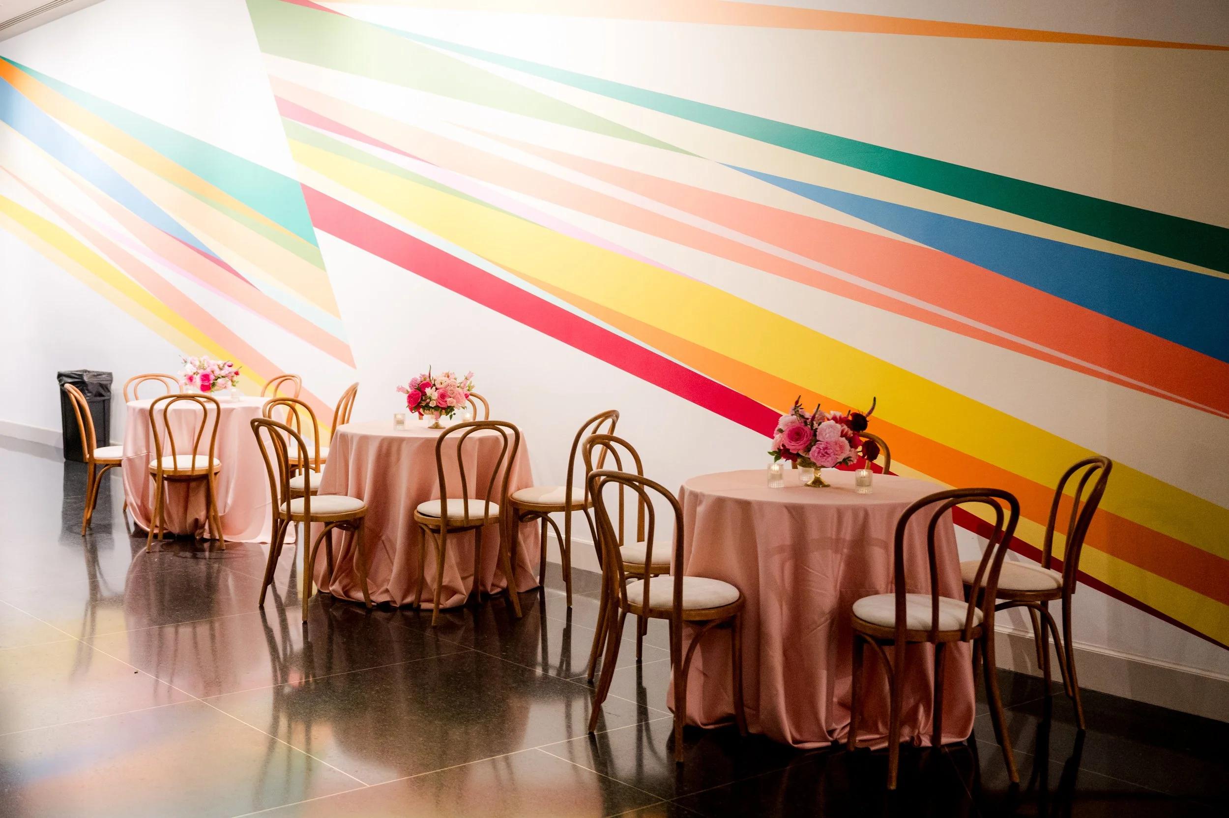 Dining area with round tables covered in pink tablecloths, each decorated with floral centerpieces, and surrounded by wooden chairs. There is a colorful, geometric mural on the white wall in the background.