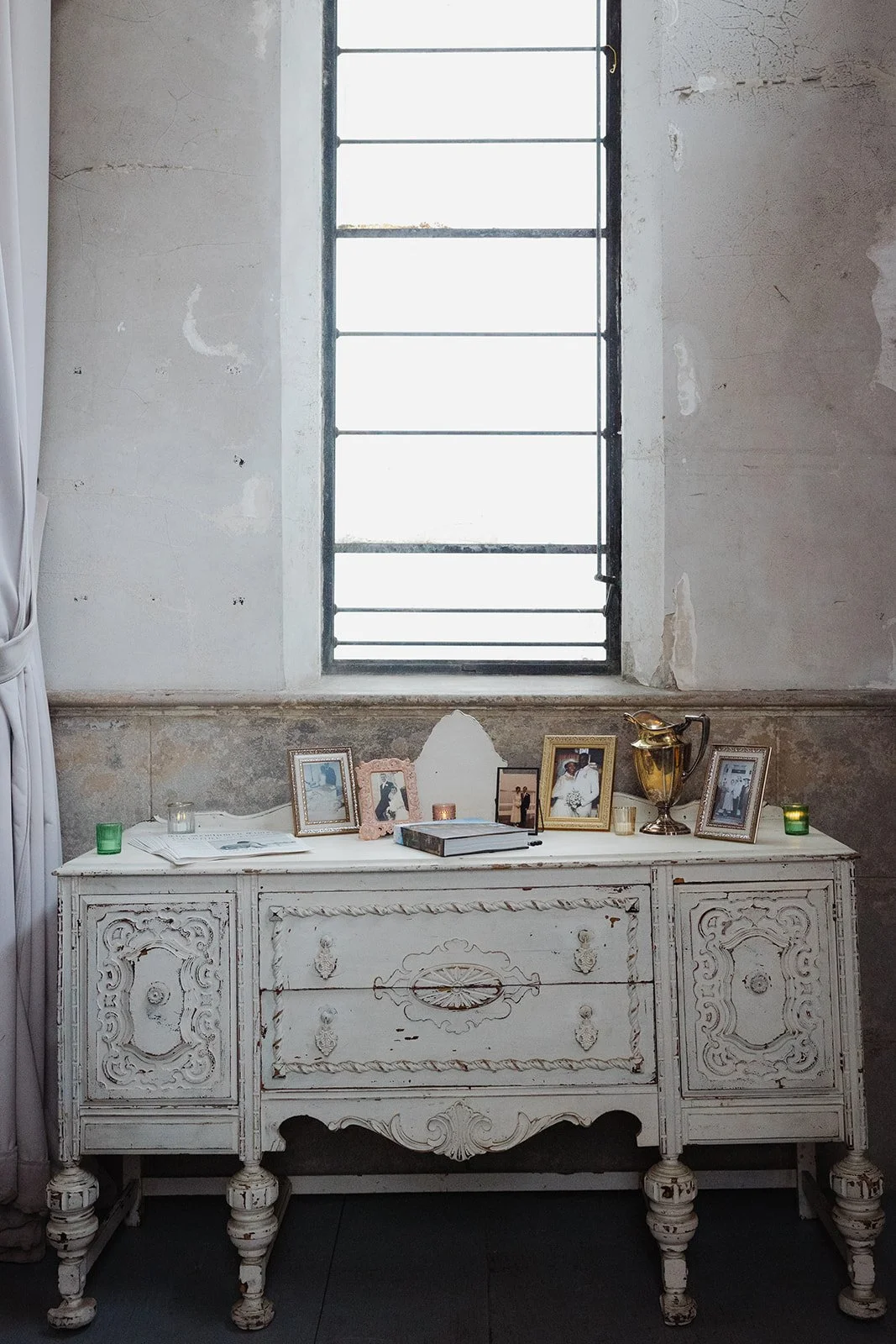 An antique white dresser with ornate carvings, placed beneath a tall, narrow window with black metal bars. The dresser is decorated with framed photographs, candles, a silver pitcher, and assorted items, set against a rustic, weathered wall.