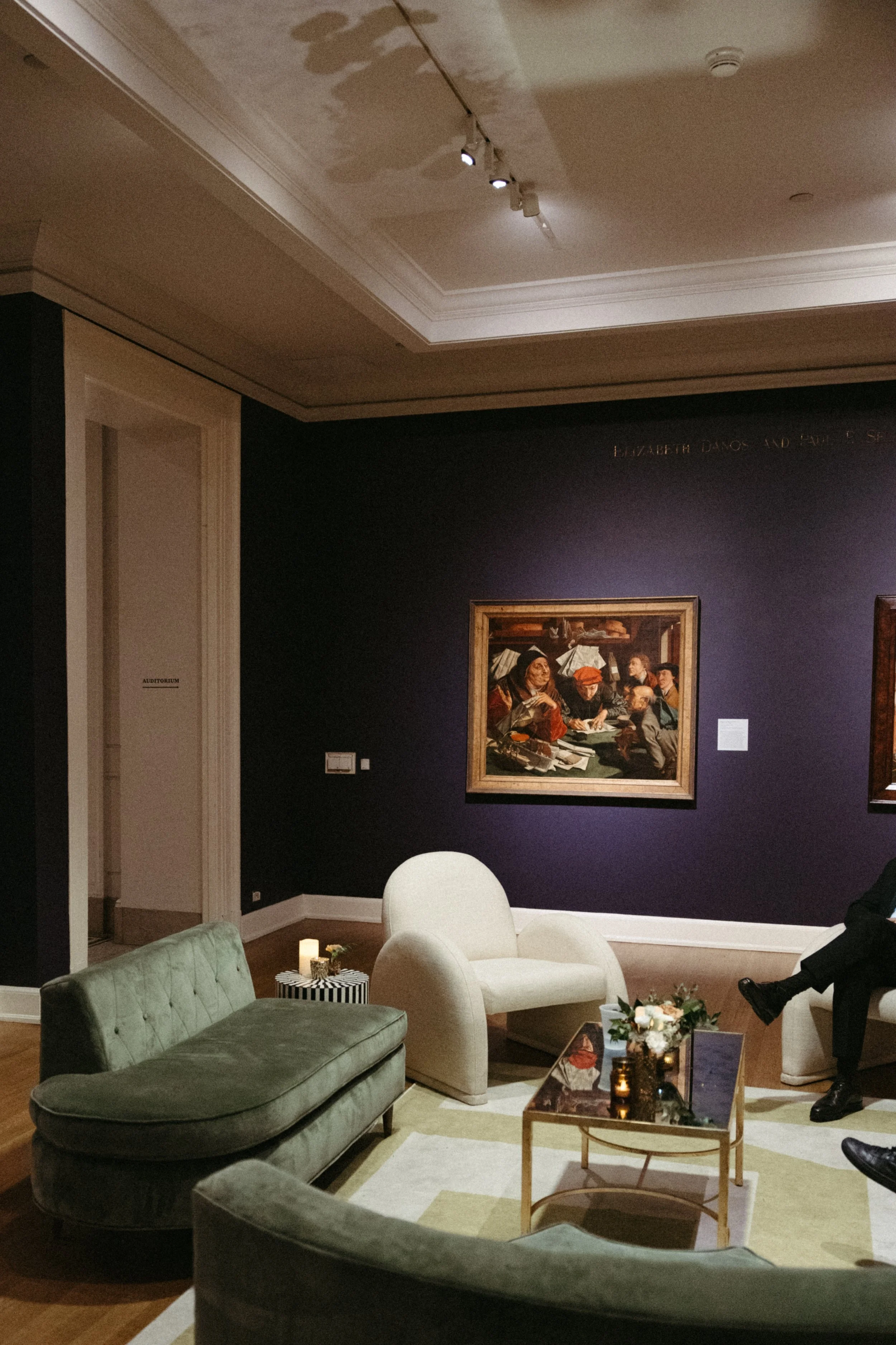 A cozy lounge area in an art gallery with a dark purple wall, paintings, and modern furniture including green and white chairs, a coffee table with flowers, candles, and decorative items, and a person seated on a white chair.