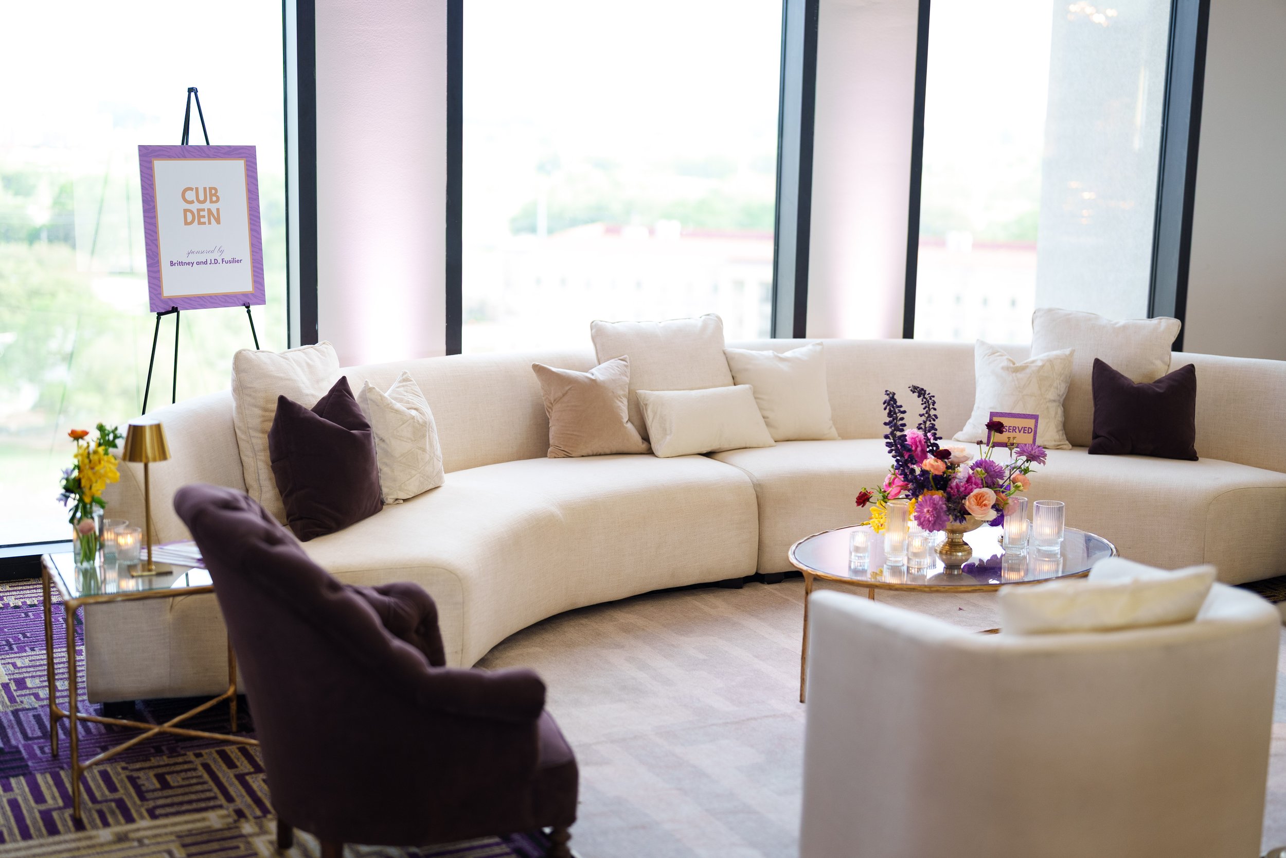 A spacious event room with large windows, a white L-shaped sofa with multiple pillows, a round coffee table with a floral centerpiece, and a sign on an easel that reads 'CUB DEN'.