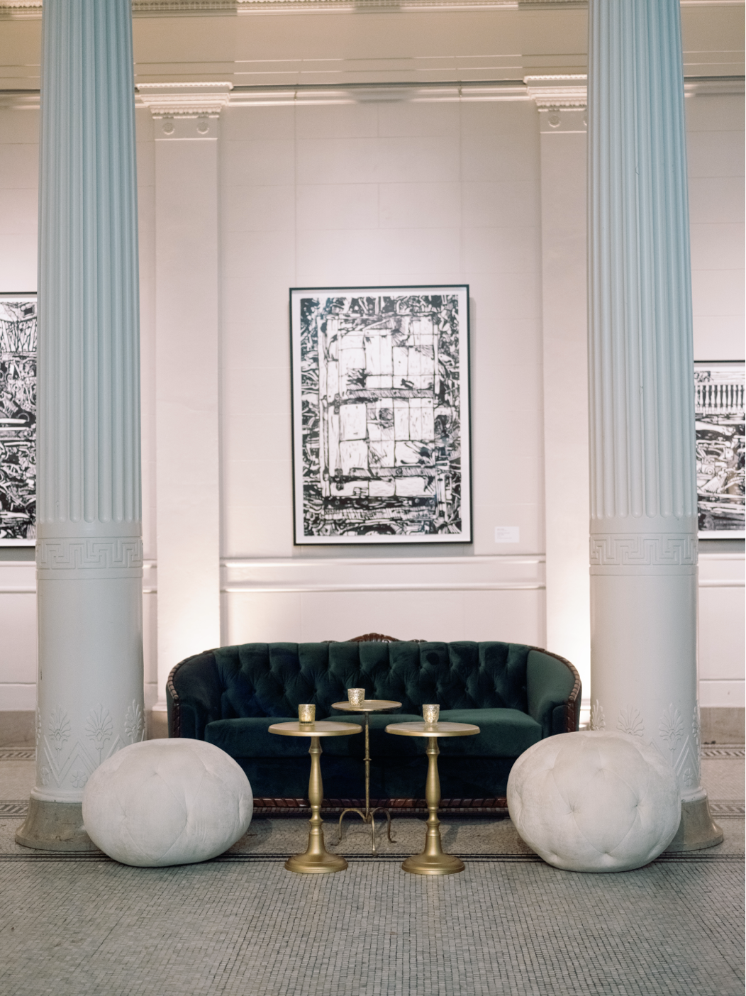Elegant hotel lobby with a dark green tufted velvet sofa, gold side tables with candles, large ornate white columns, abstract black and white artwork on the wall, and plush white poufs on a patterned carpet.