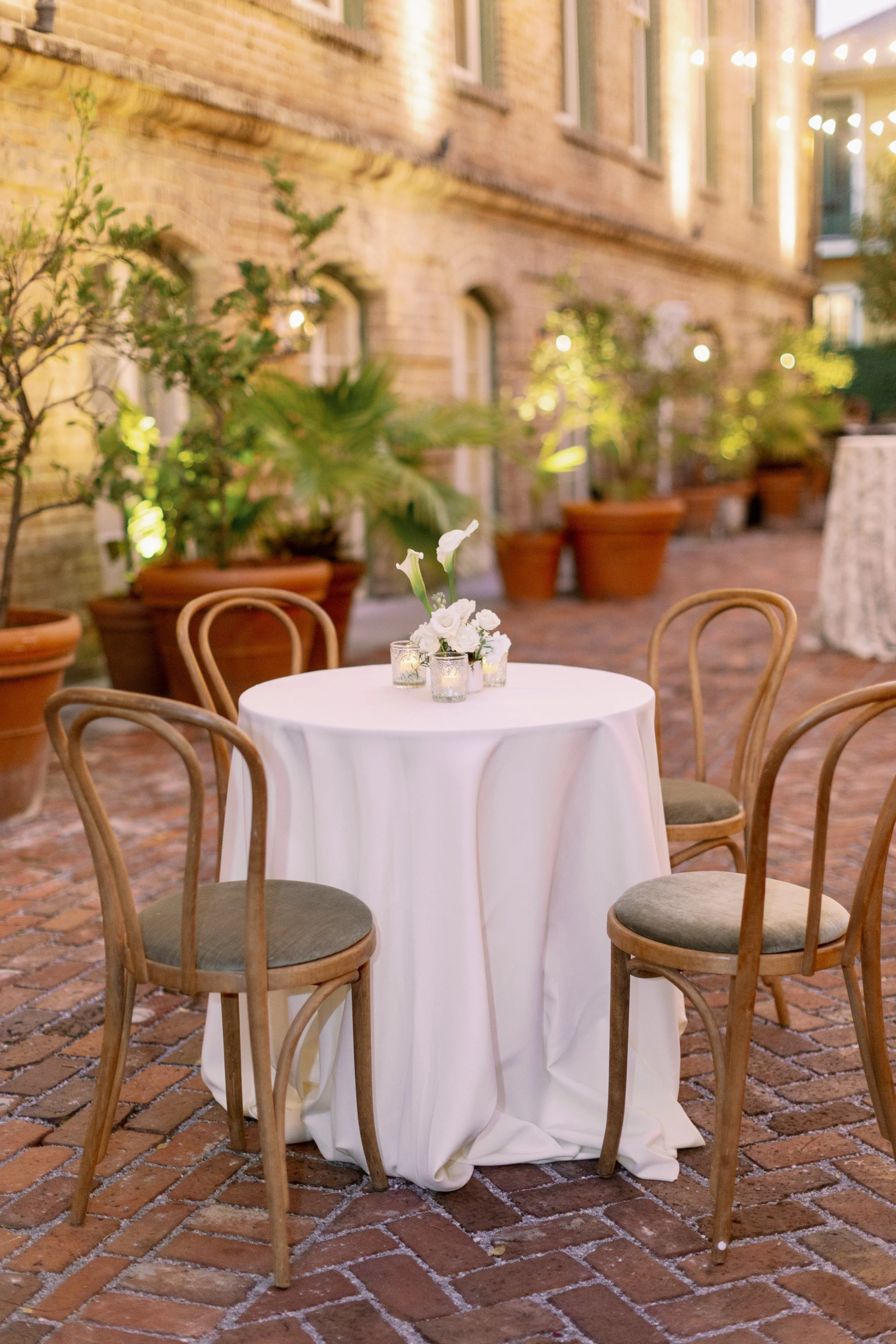Outdoor event setup with a round table covered in a white tablecloth, decorated with a floral centerpiece and candles. Surrounding the table are wooden chairs with cushioned seats. Potted plants and string lights are visible in the background, with a
