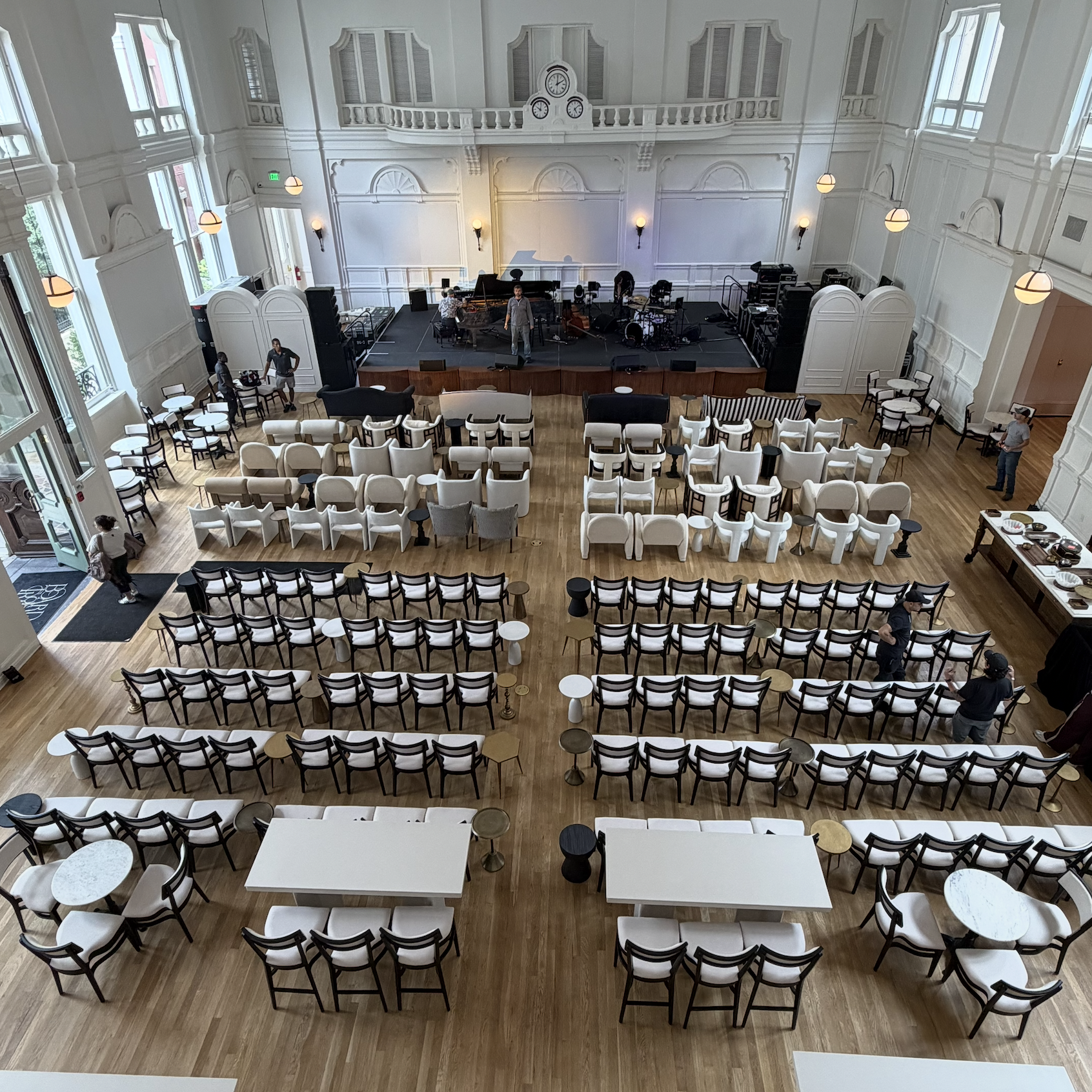 Concert hall with a stage and chairs arranged for an audience, musicians preparing to perform, and large windows allowing natural light to illuminate the room.