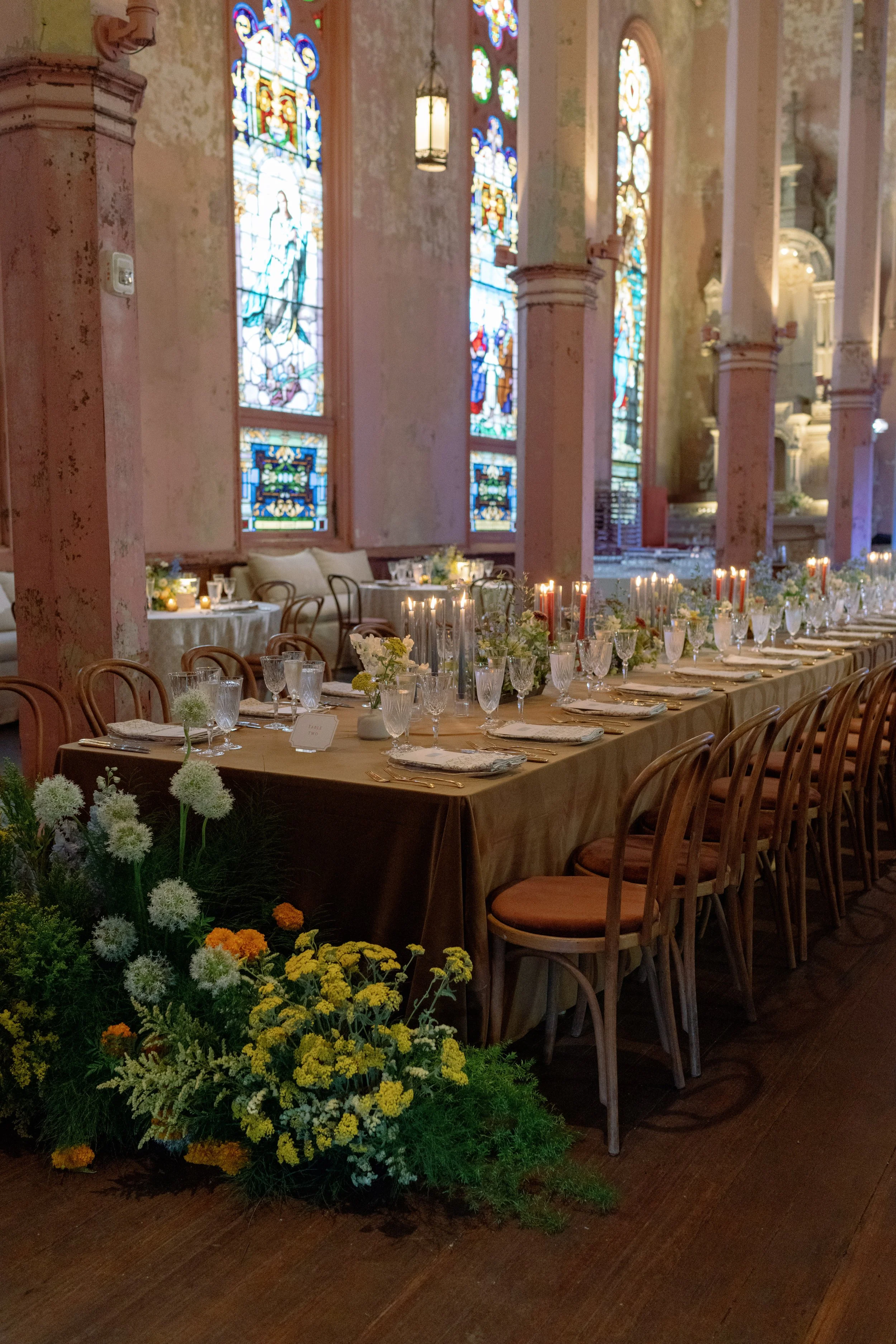 Elegant banquet table set with candles, glassware, plates, and floral centerpieces inside a church with stained glass windows and pink columns.