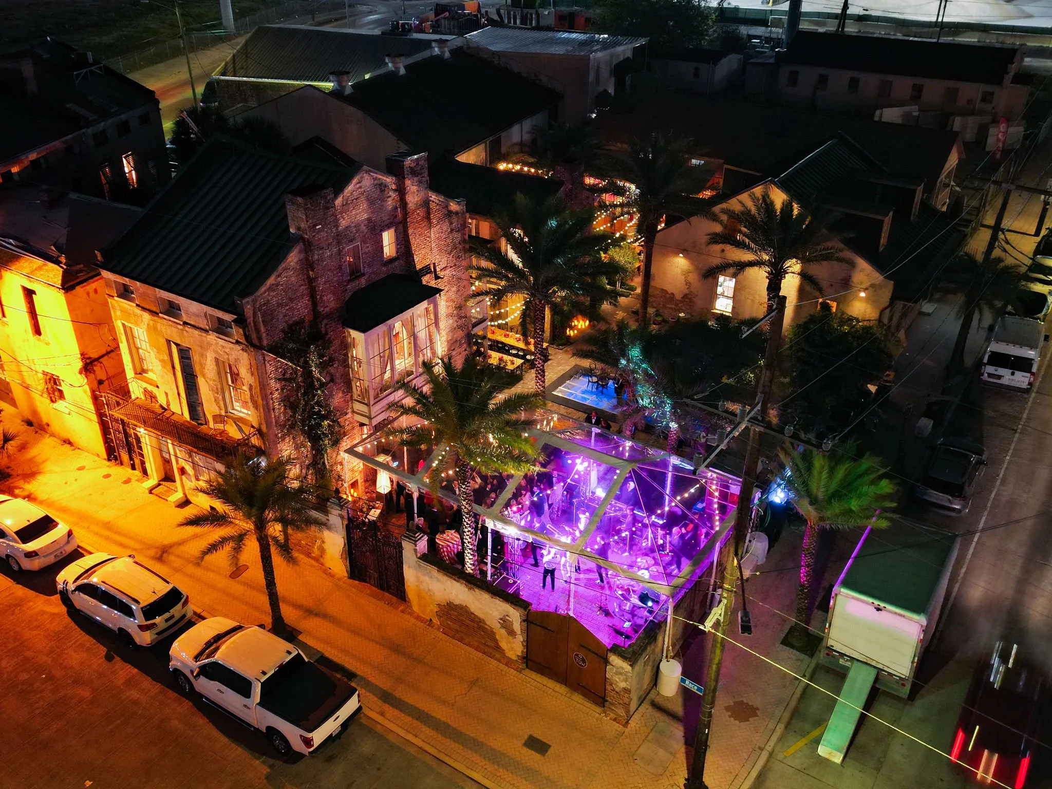 Nighttime aerial view of a building with outdoor lighting, trees, and a covered patio area visible. Several parked cars are on the street.