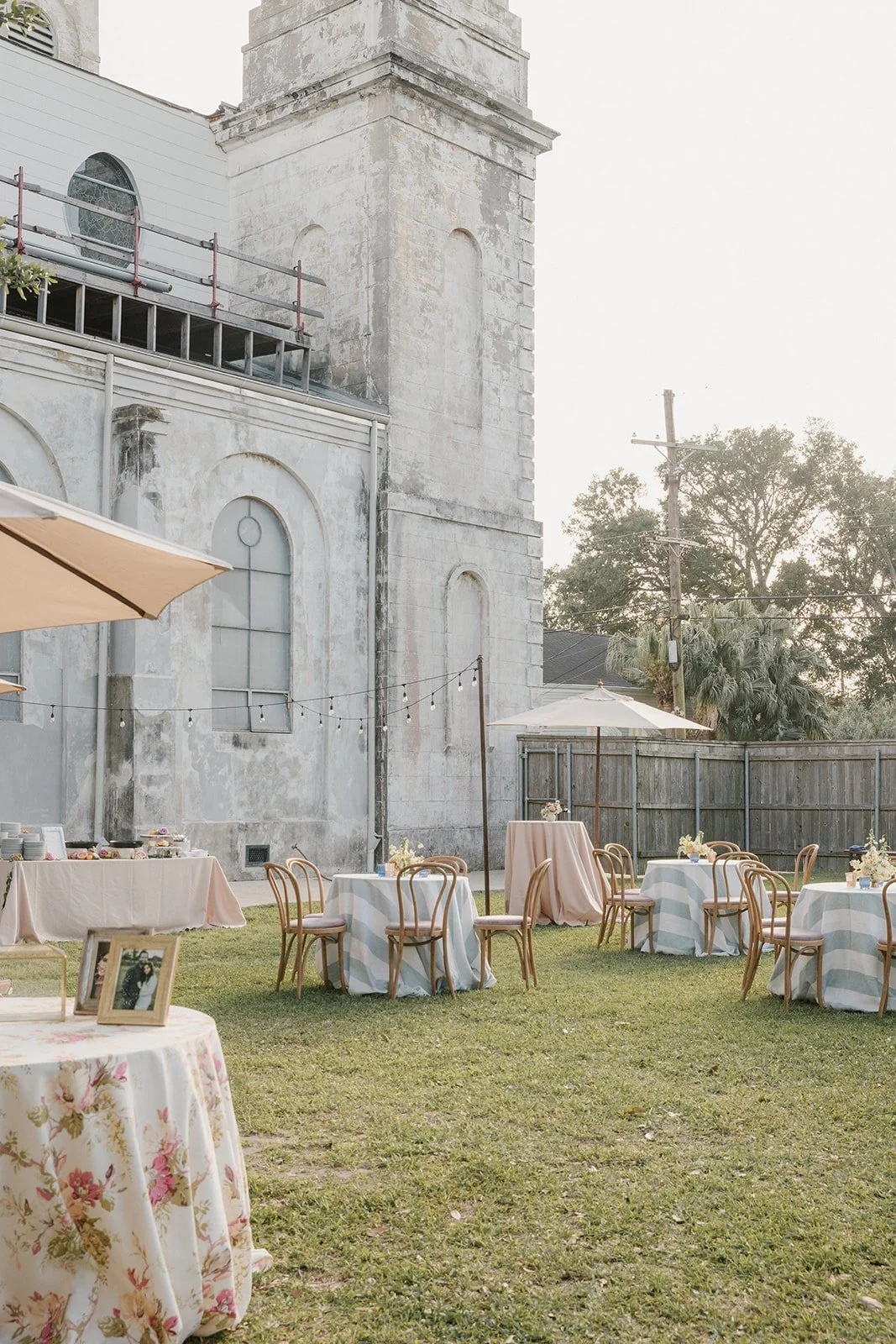 Outdoor event setup with round tables, chairs, umbrellas, and string lights, near an old stone building with a fenced yard and trees in the background.