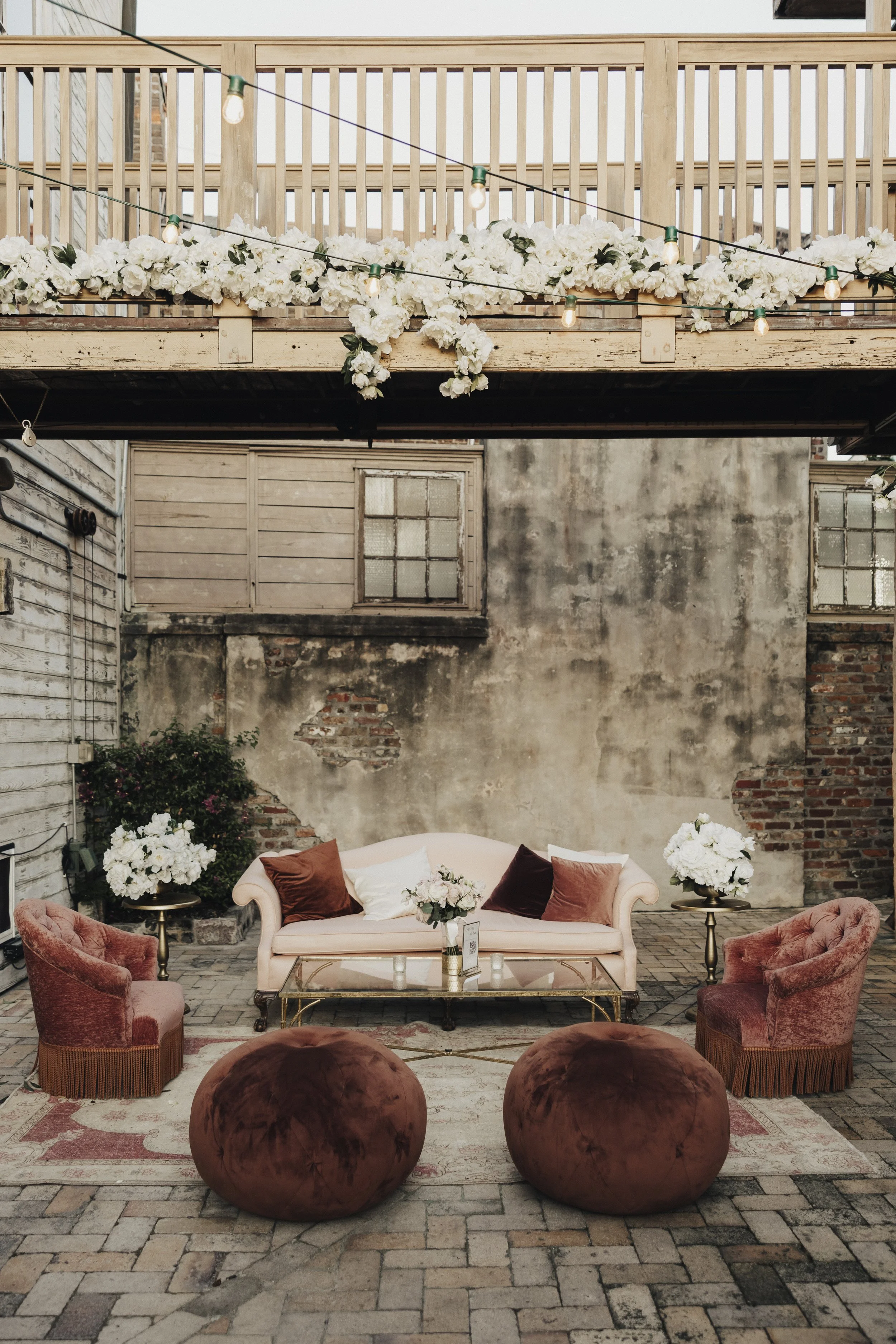 An outdoor patio decorated for a wedding or party with a pink vintage sofa, velvet chairs, floral arrangements, and string lights. There is a rustic brick and concrete wall backdrop.