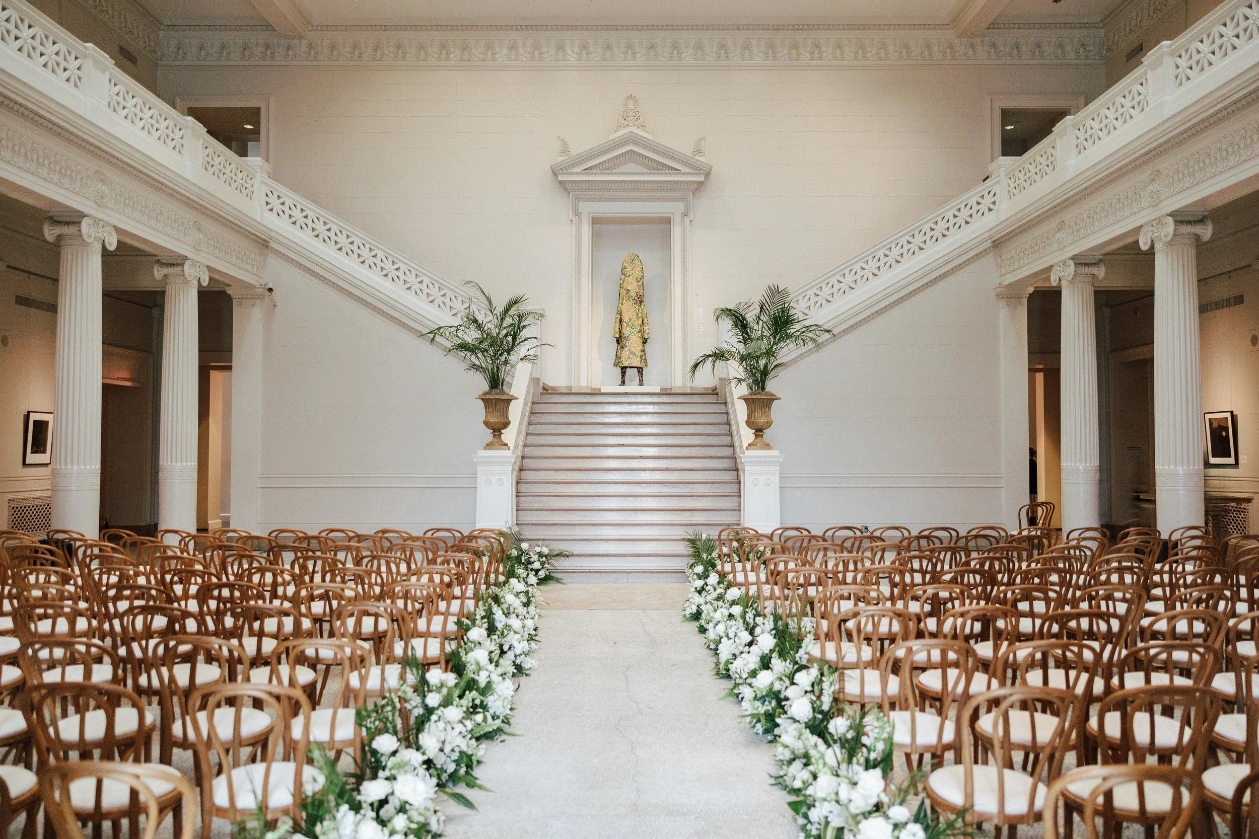 Elegant indoor wedding ceremony setup with rows of wooden chairs adorned with white flowers lining an aisle leading to a staircase. The staircase has potted plants on either side and a person in a colorful dress is at the top, in front of a classical