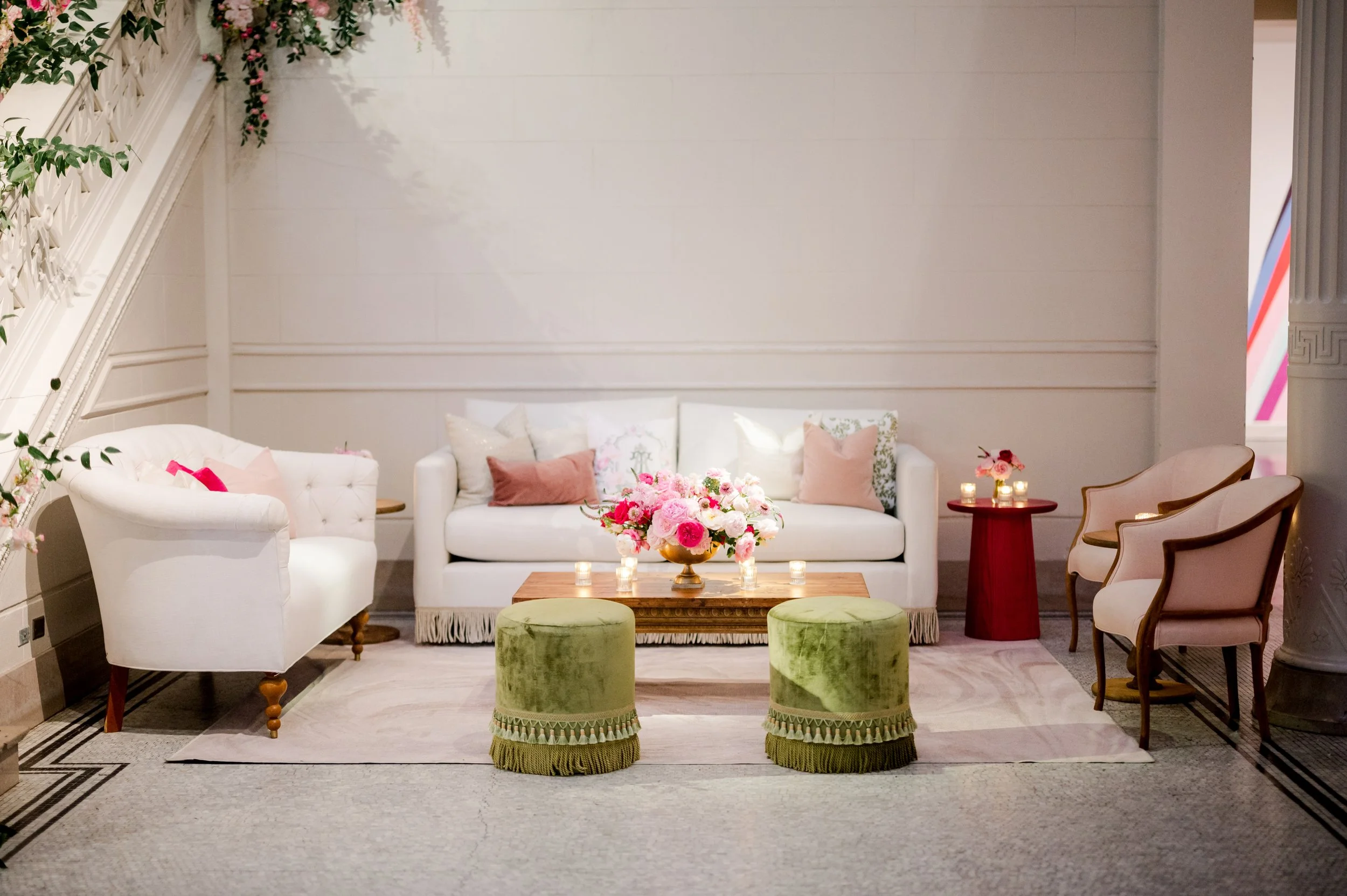 Interior of a decorated reception or lounge area with white couches, pink flowers, candles, and green ottomans.