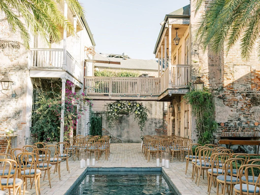 Outdoor courtyard wedding ceremony setup with chairs arranged on either side of a small pool, surrounded by rustic brick and wooden buildings with greenery and flowers.