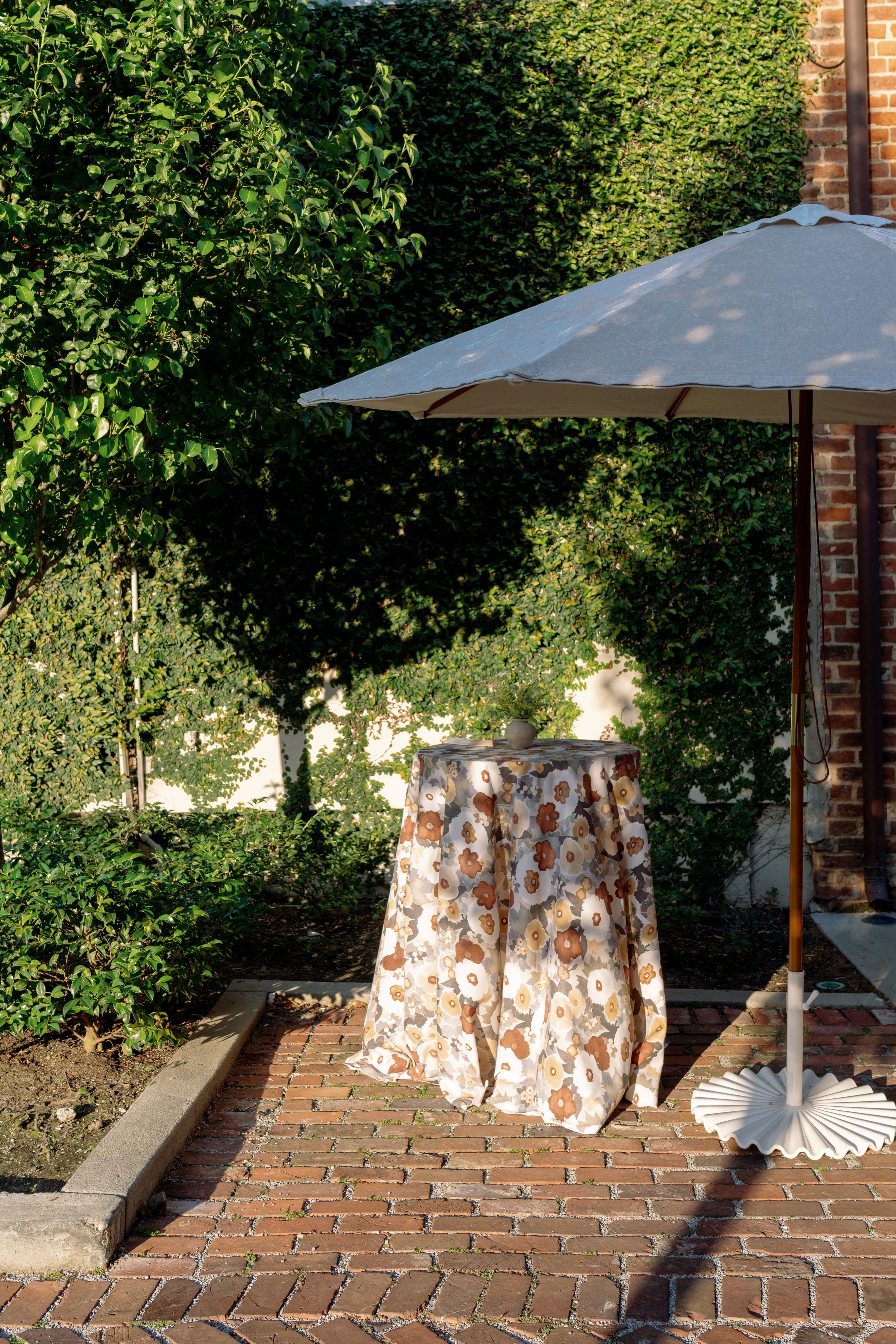 A round table covered with a floral tablecloth, positioned on a brick patio, with a large beige patio umbrella shading it. Green bushes and trees are in the background, and a brick building wall is partially visible on the right side.
