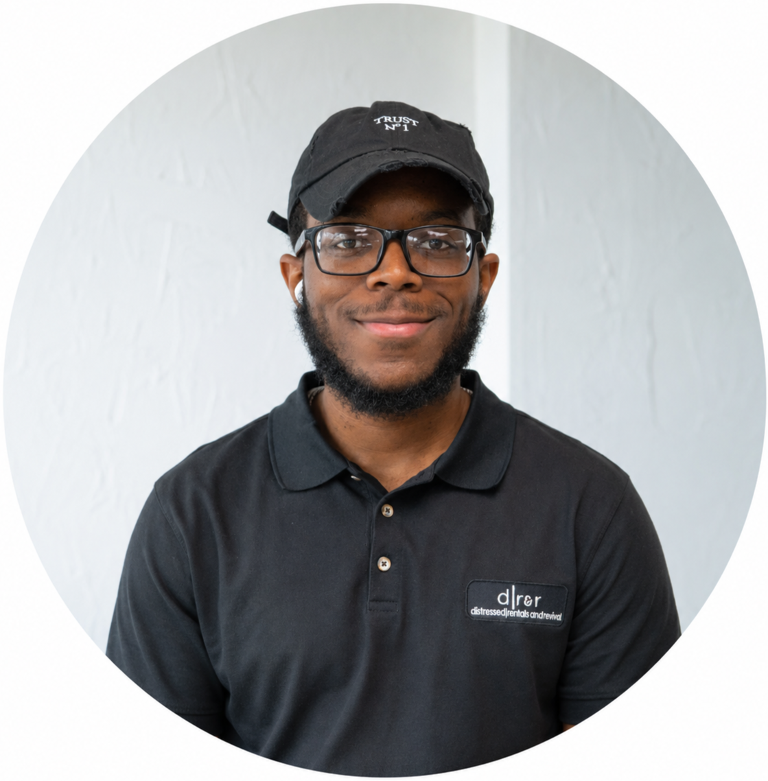 A young man with glasses, a beard, and wearing a black cap and black polo shirt, smiling and standing against a white wall.