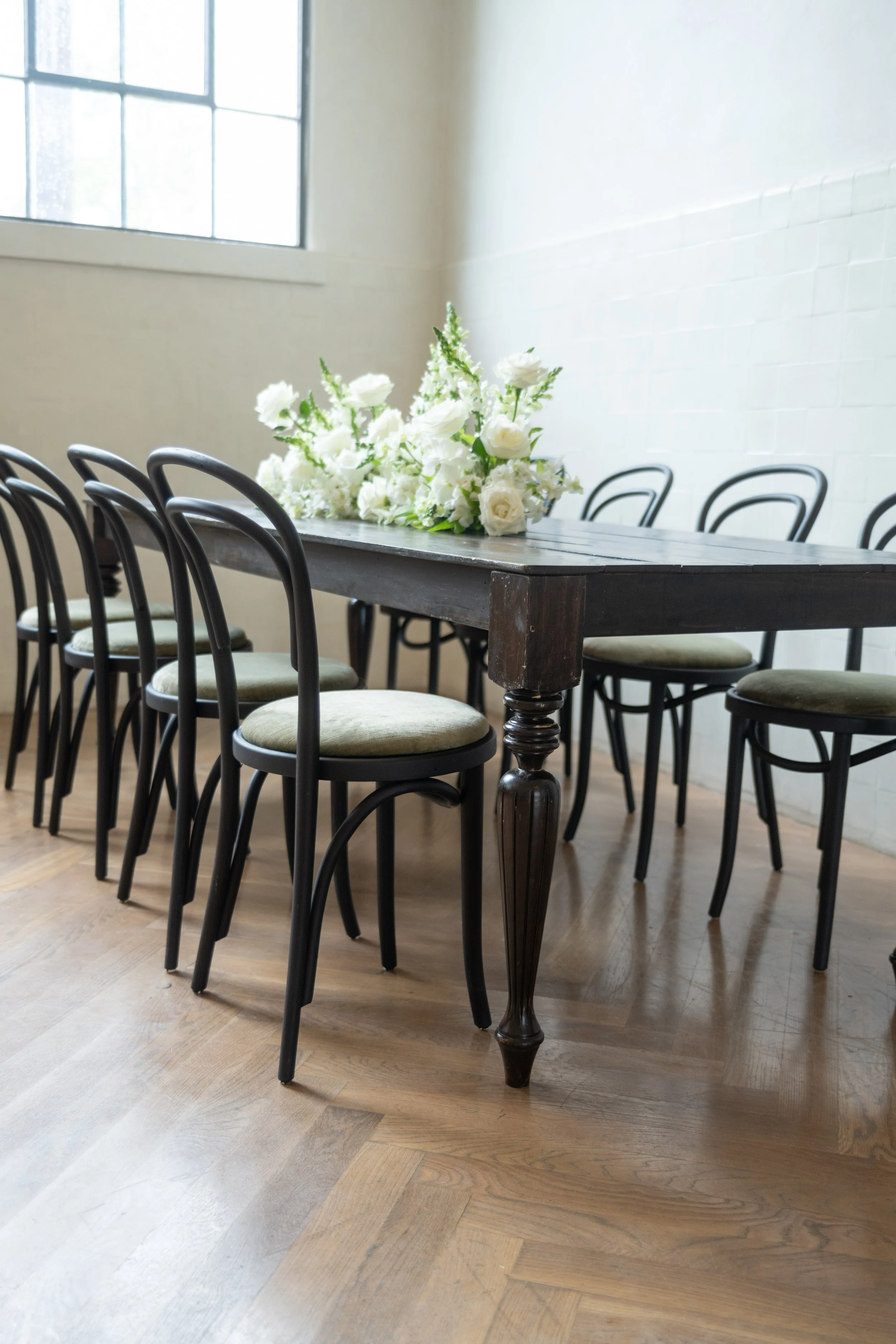 A dark wooden dining table with a floral centerpiece of white flowers and green leaves, surrounded by black chairs with beige cushions, in a room with a large window and white walls.