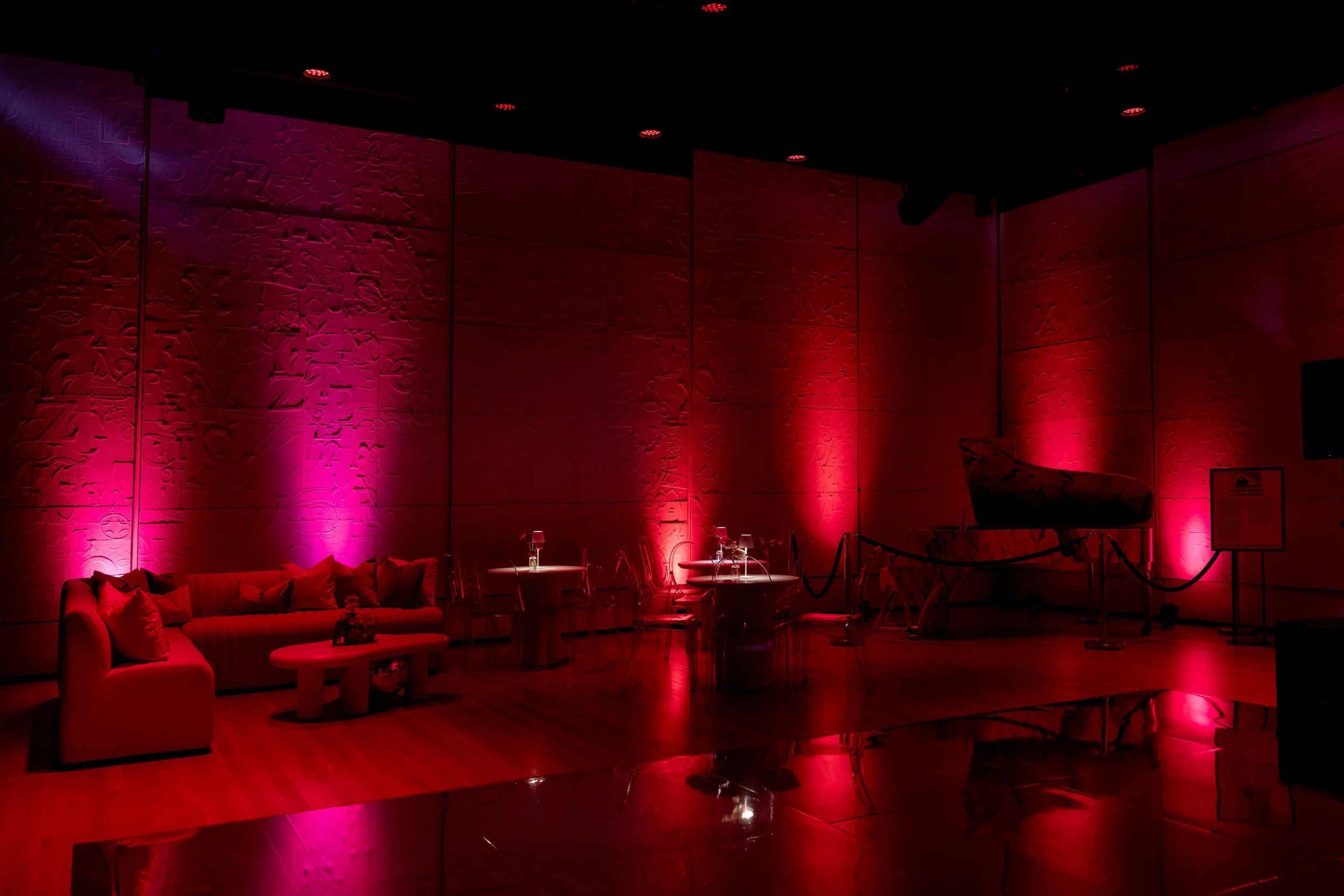 Interior of a dimly lit room with red and purple lighting, decorated with abstract wall art, and furnished with a sofa, chairs, and tables, possibly for a lounge or event space.