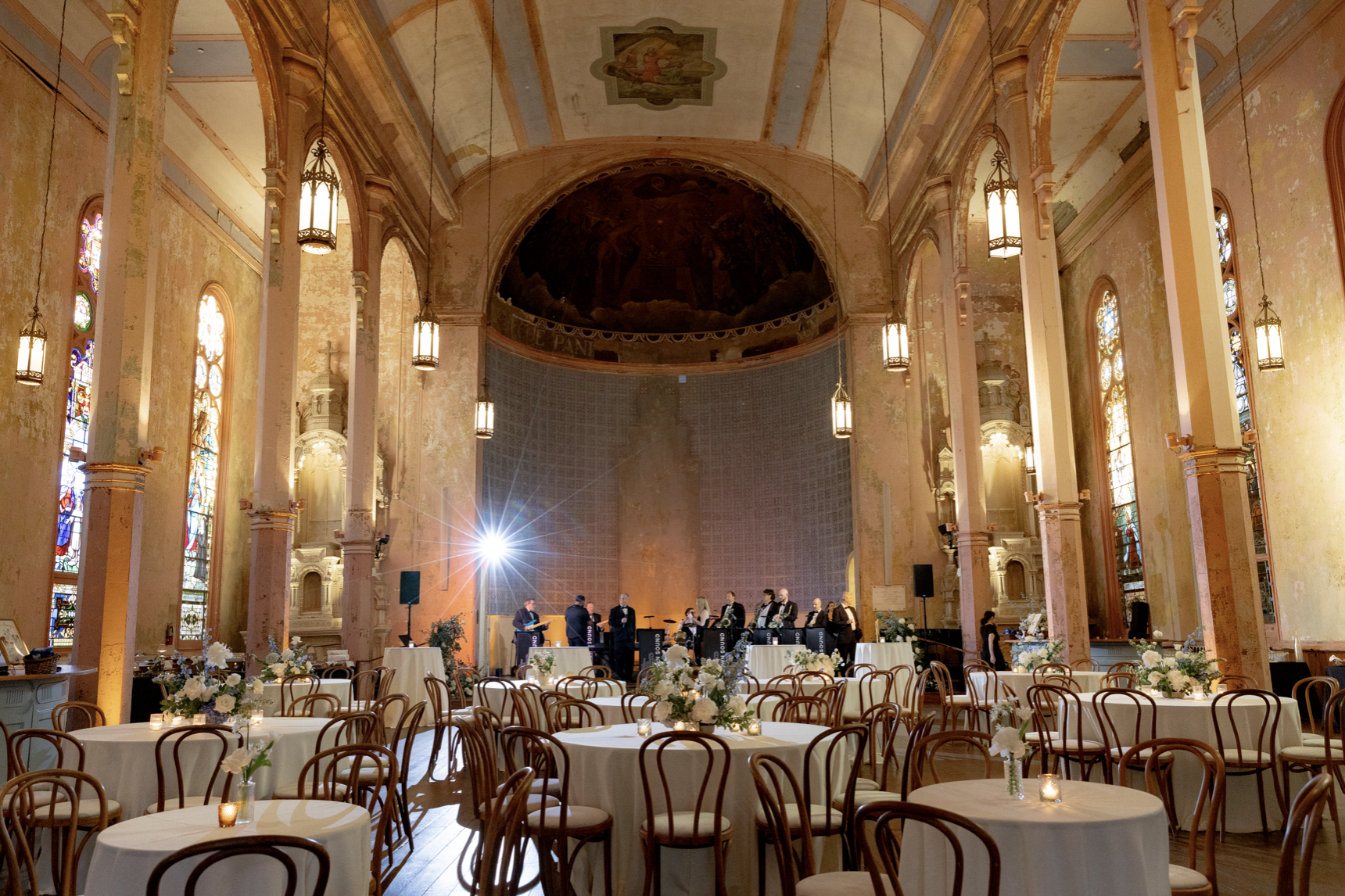 Inside a church decorated for a wedding with round tables and floral centerpieces, a band is performing on stage, stained glass windows line the walls, and chandeliers hang from the high ceiling.