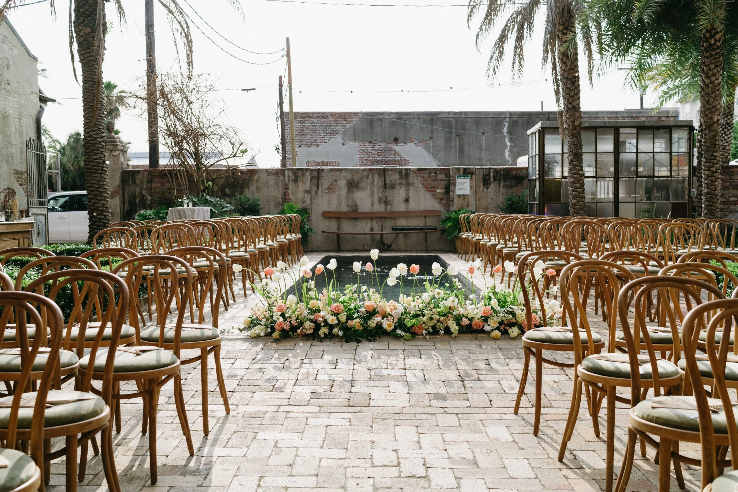Outdoor venue prepared for a wedding ceremony with rows of wooden chairs and a floral arrangement at the front, palm trees, a small water feature, and an unfinished brick wall in the background.