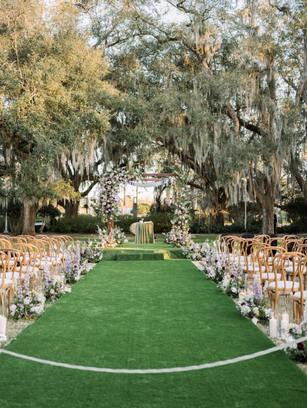Outdoor wedding ceremony setup with an aisle of green grass, wooden chairs on either side, and floral arrangements with purple, pink, and white flowers at the entrance of an arch under large oak trees draped with Spanish moss.