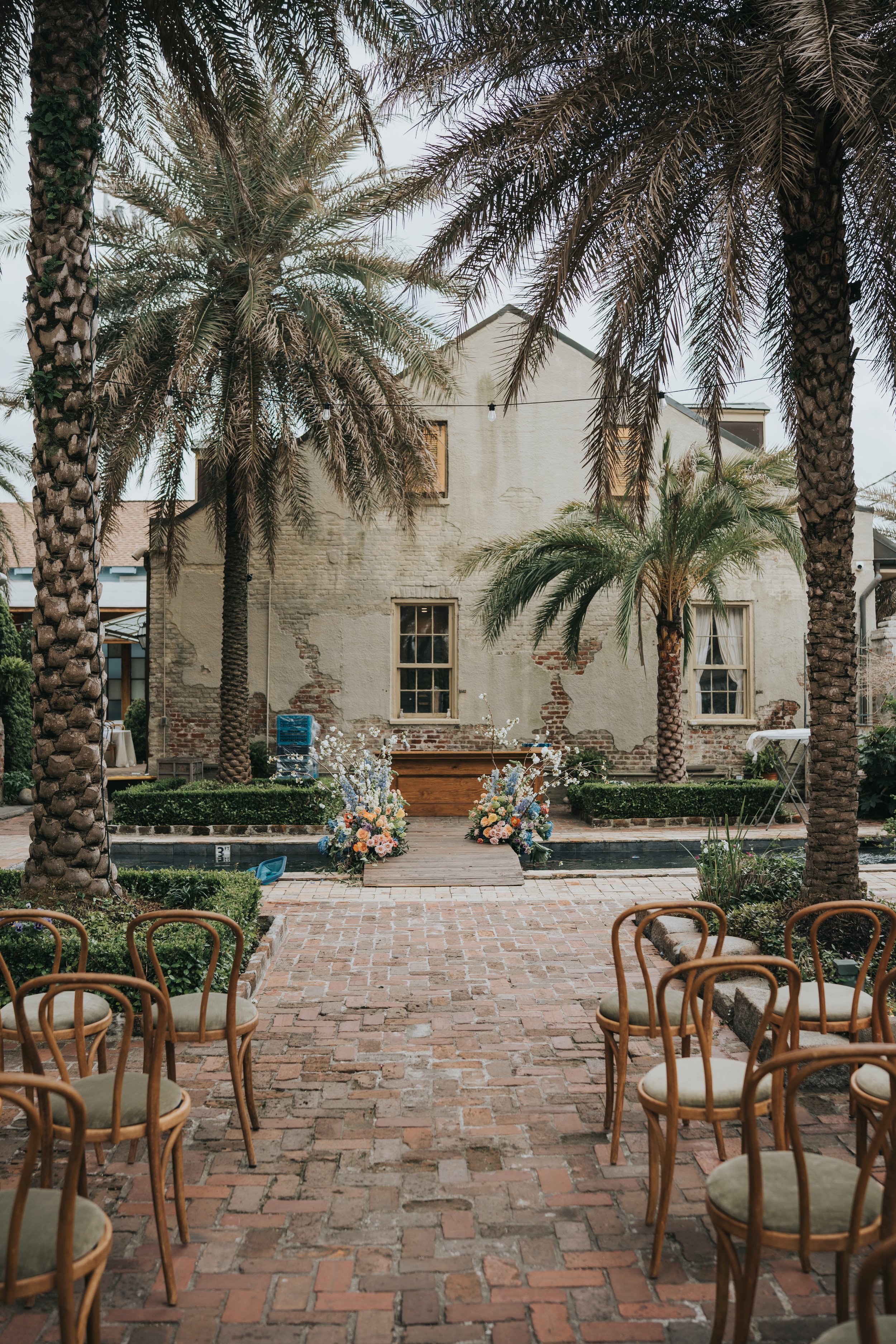 Outdoor wedding setup with rows of chairs, brick pathway, floral arrangements, palm trees, and a rustic building in the background.