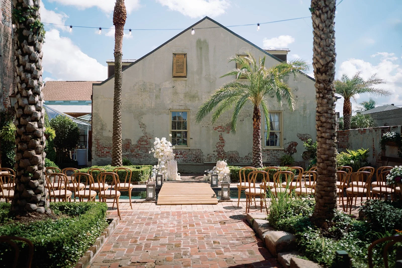 Outdoor wedding setup with chairs arranged on both sides of a brick pathway leading to a small wooden platform, decorated with white flowers and lanterns, surrounded by green plants and tall palm trees in a courtyard with an aged brick and stucco wal