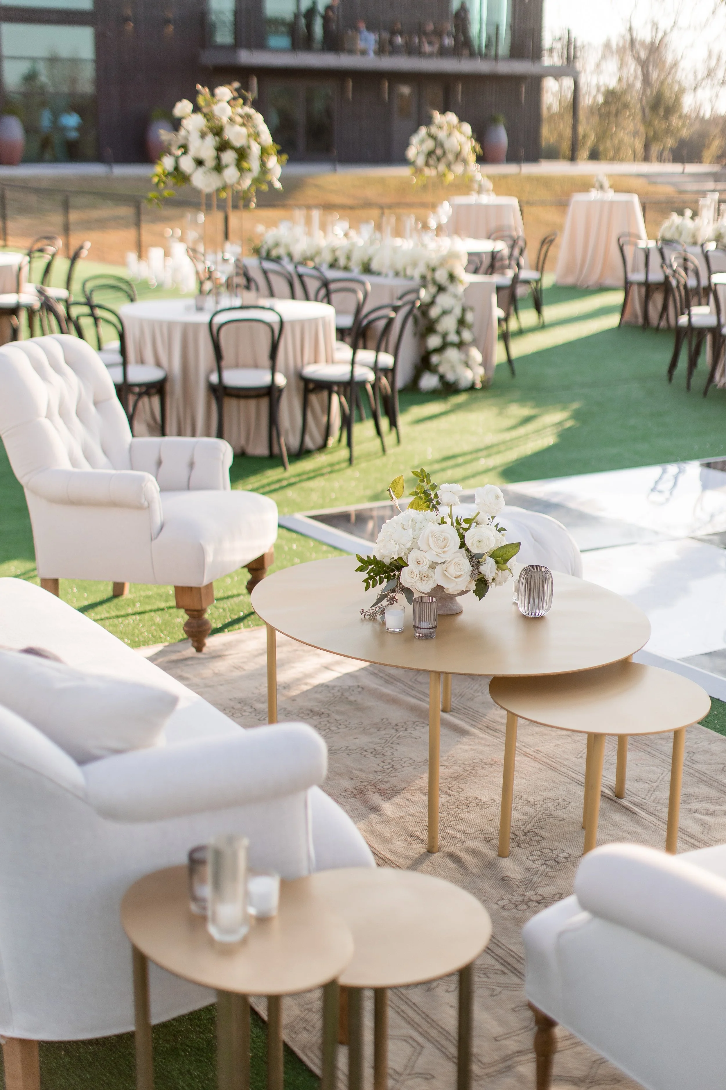 Elegant outdoor wedding reception with white upholstered chairs, round tables with beige tablecloths, and floral centerpieces, set on a grassy area with a modern building in the background.