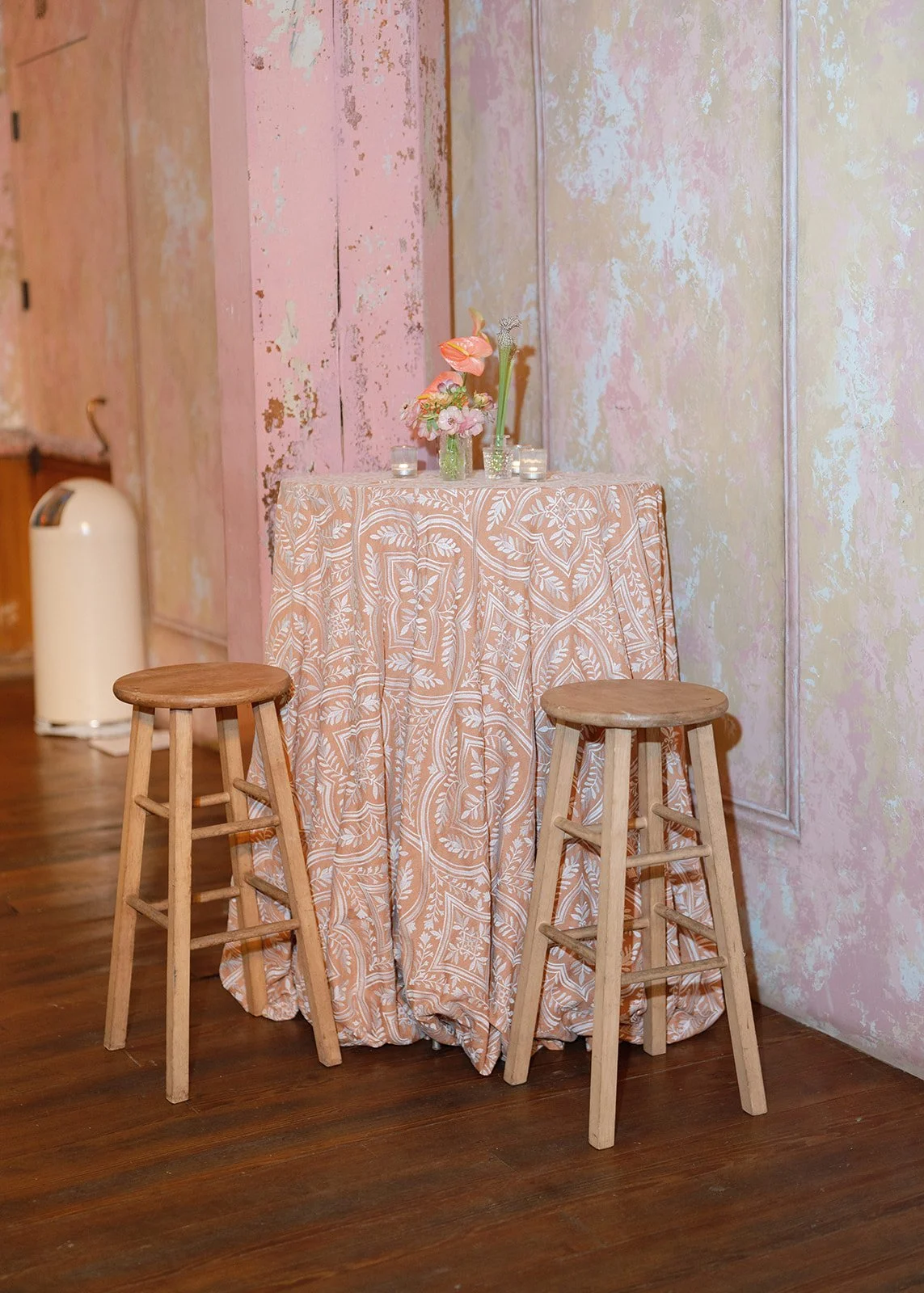 A tall table with a pink and white patterned tablecloth, decorated with pink and peach flowers in a vase and small candles. Two wooden stools are placed in front of the table. The background has textured pink and yellow walls, and to the left, there'