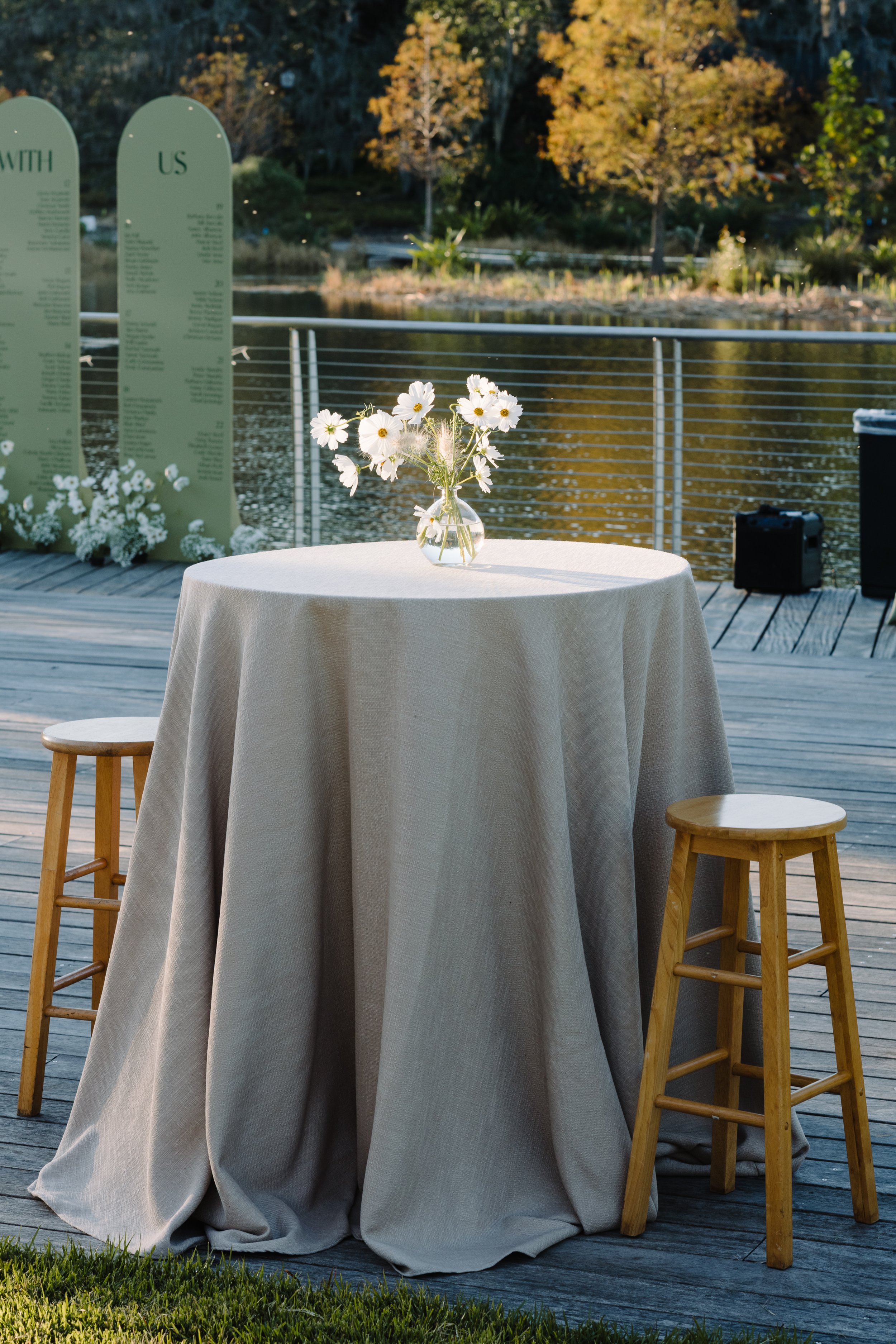 A round table with a beige tablecloth, a vase with white flowers, wooden stools, a lakeside setting with trees, and memorial plaques in the background.