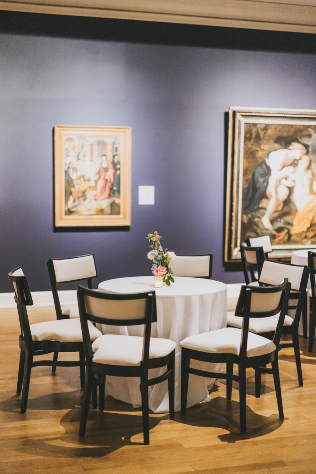 A round table with a white tablecloth and a floral centerpiece, surrounded by six black and white chairs, in an art gallery with two large paintings on a purple wall.