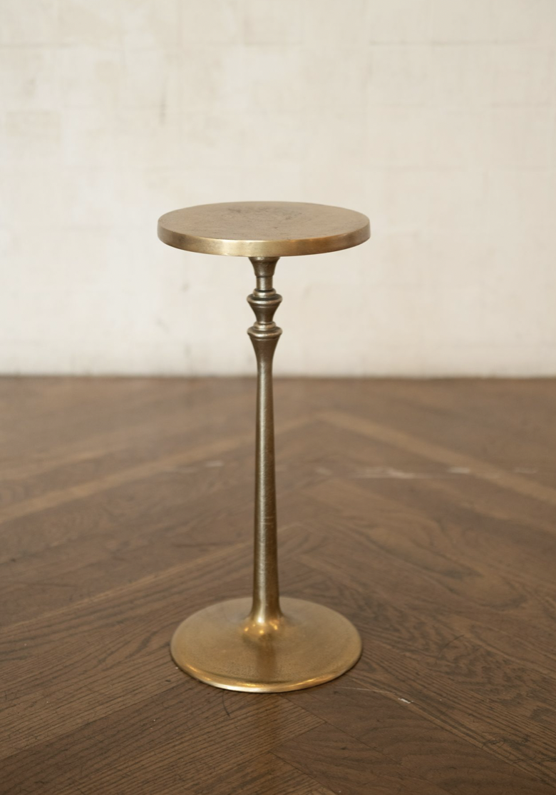 A tall, thin, brass-colored candle holder with a round, flat top and a wide round base, standing on a wooden surface against a plain, light-colored background.