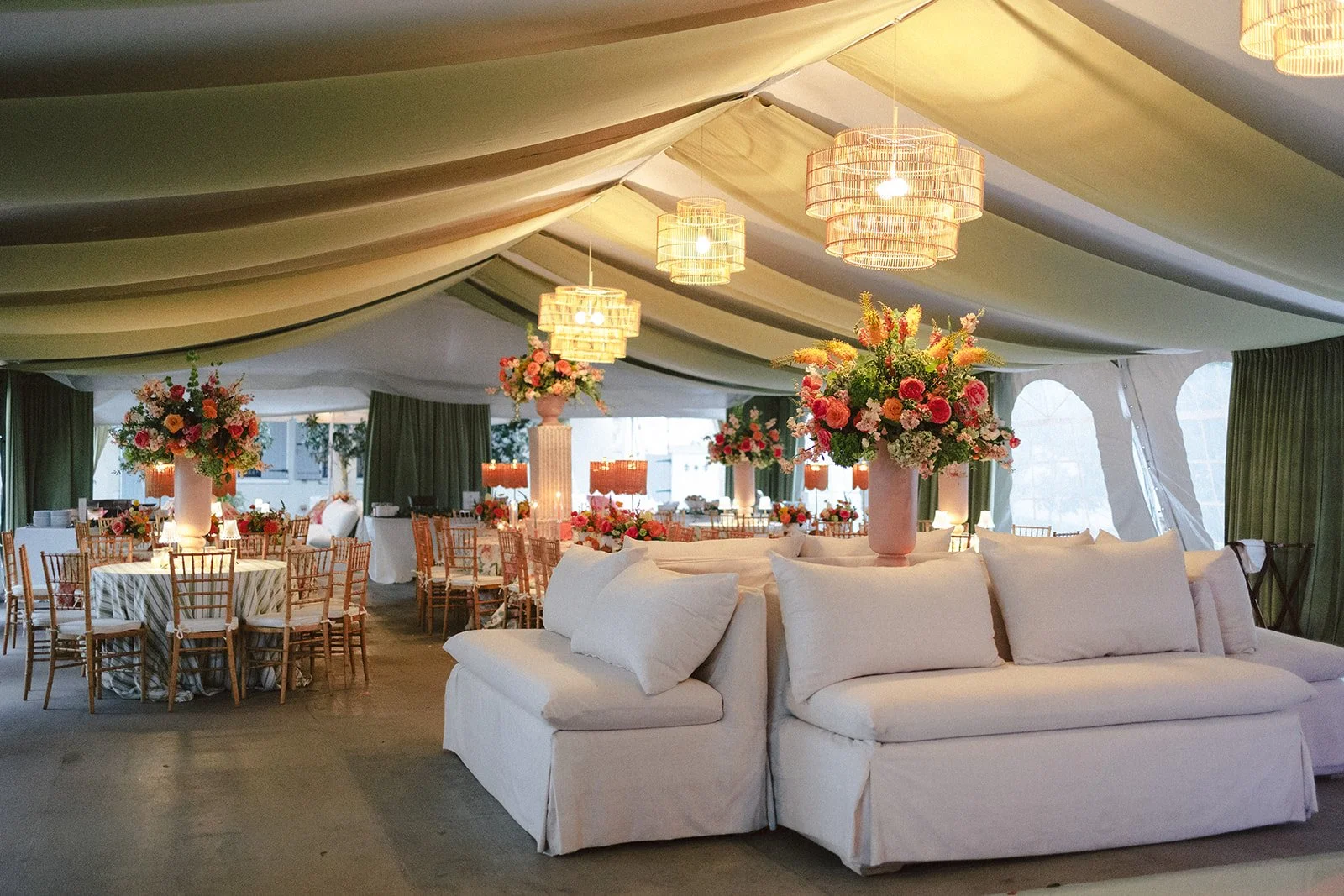 Interior of a decorated event space with a white sofa, large floral arrangements in pink vases, round tables with striped tablecloths, wooden chairs, and hanging wicker light fixtures under a draped ceiling with green curtains and large windows in th