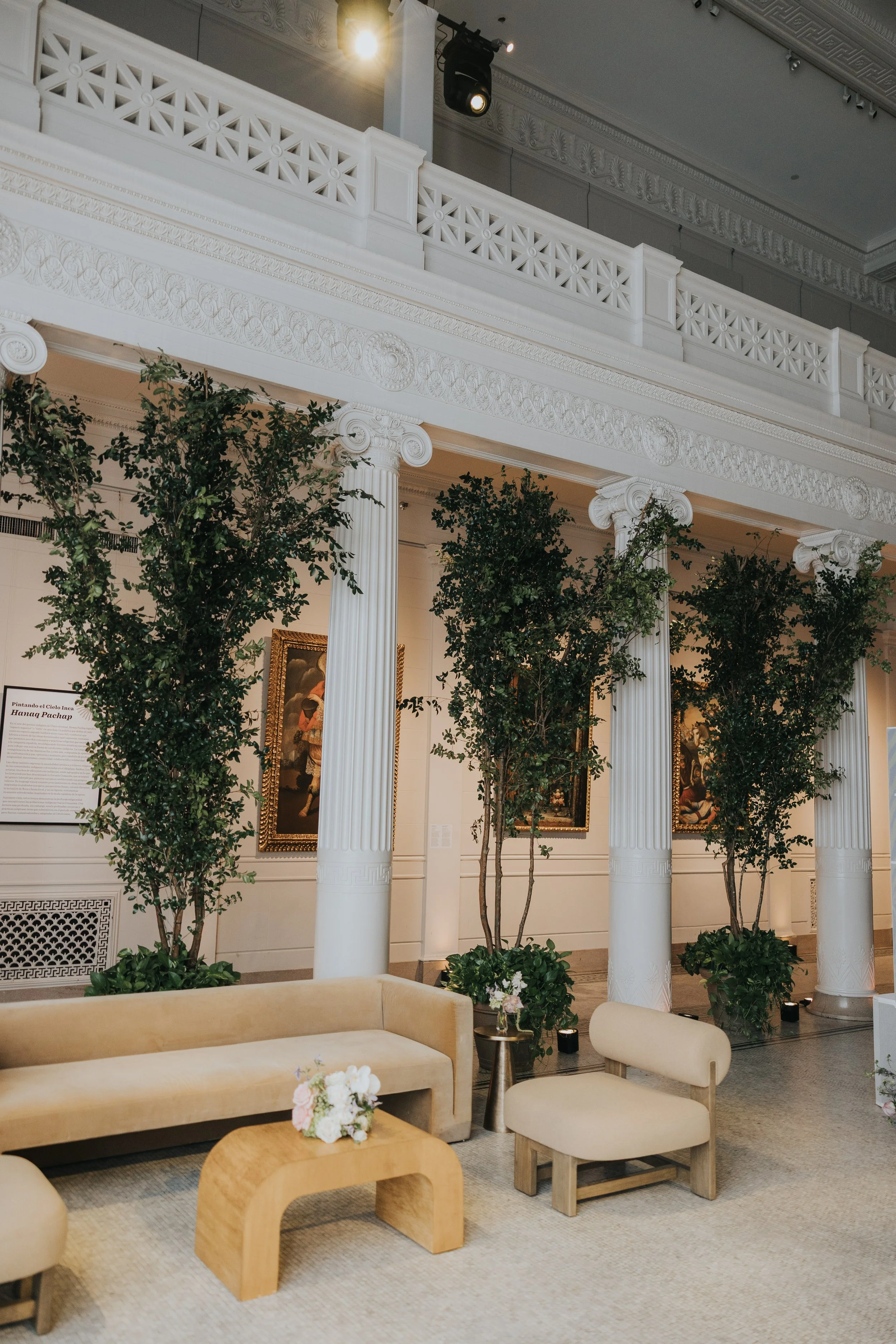 Elegant interior with white classical columns, greenery, and framed artwork, featuring beige seating and a small flower arrangement on a wooden coffee table.