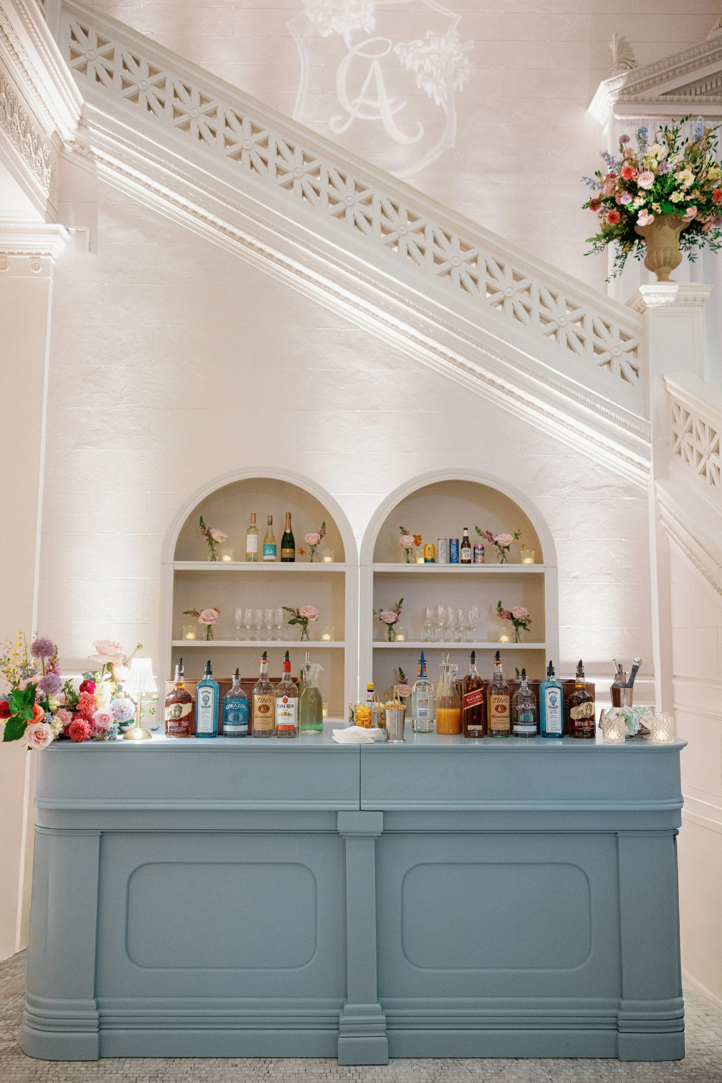 A bar setup with bottles of alcohol, cocktail shakers, and glasses on a blue counter. Behind the bar, there are two white shelves with flowers and bottles, with a decorative alcove. The room has ornate white crown molding and a chandelier's shadow on
