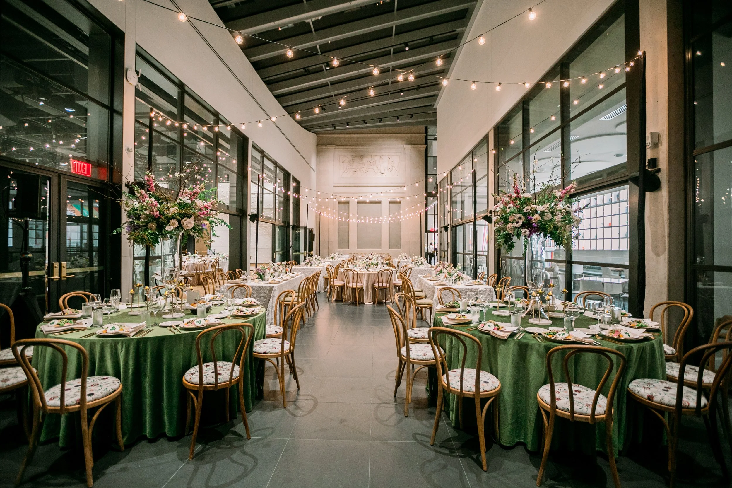 The image shows a decorated event space with large windows, string lights, and floral arrangements. There are round tables with green tablecloths and floral-patterned chairs, set with plates, glasses, and napkins, ready for a celebration or banquet e