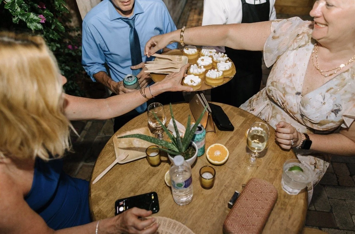 Group of people celebrating around a table with cupcakes, drinks, and a potted plant.