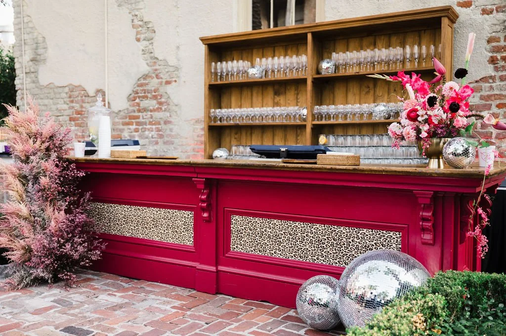 A bar or counter with a deep red finish and leopard print panels, decorated with pink flowers, pink foliage, and disco balls, set against a brick wall with a wooden shelf holding glasses.