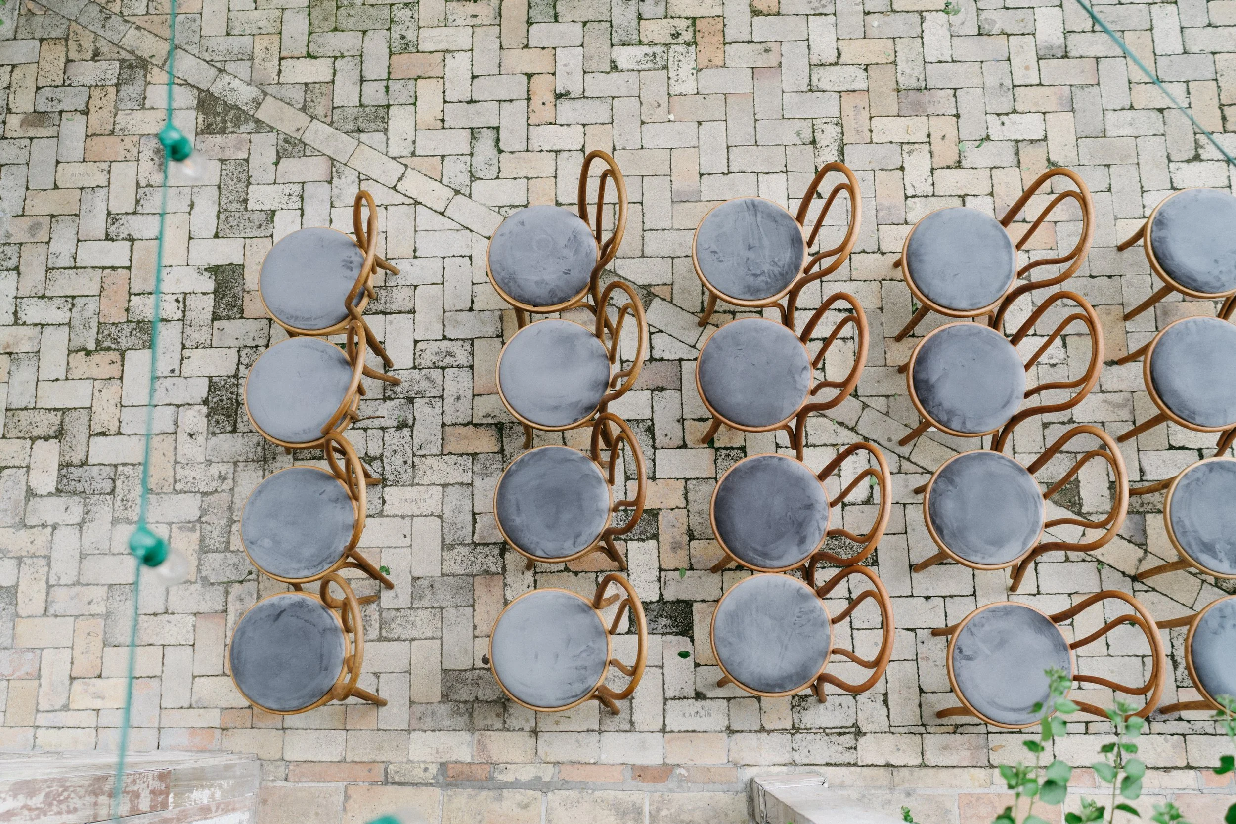 A top-down view of several round tables with gray tops and wooden chairs arranged around them on a stone-tiled outdoor patio.