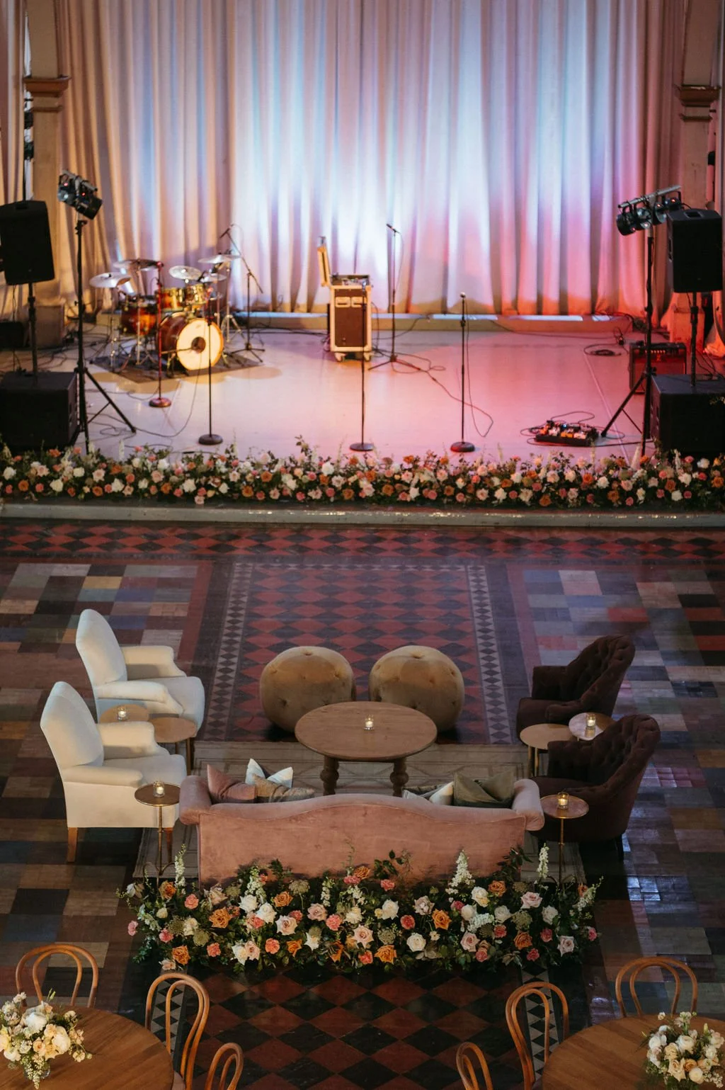 Stage with musical instruments, microphones, and lighting, decorated with flowers, in front of a seating area with chairs, sofas, and tables adorned with candles, in a banquet hall.