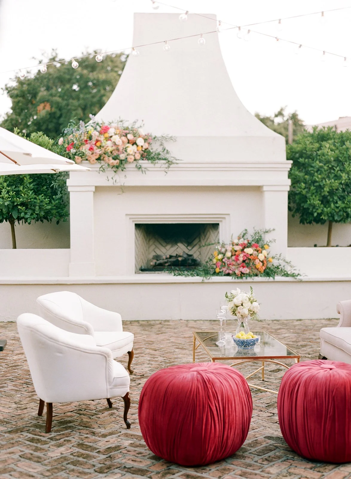 Outdoor patio with white fireplace decorated with pink and white flowers, white and red upholstered seating, a glass coffee table with a floral arrangement, two wine glasses, and a bowl of lemons. String lights are hanging overhead, greenery surrounds the area.
