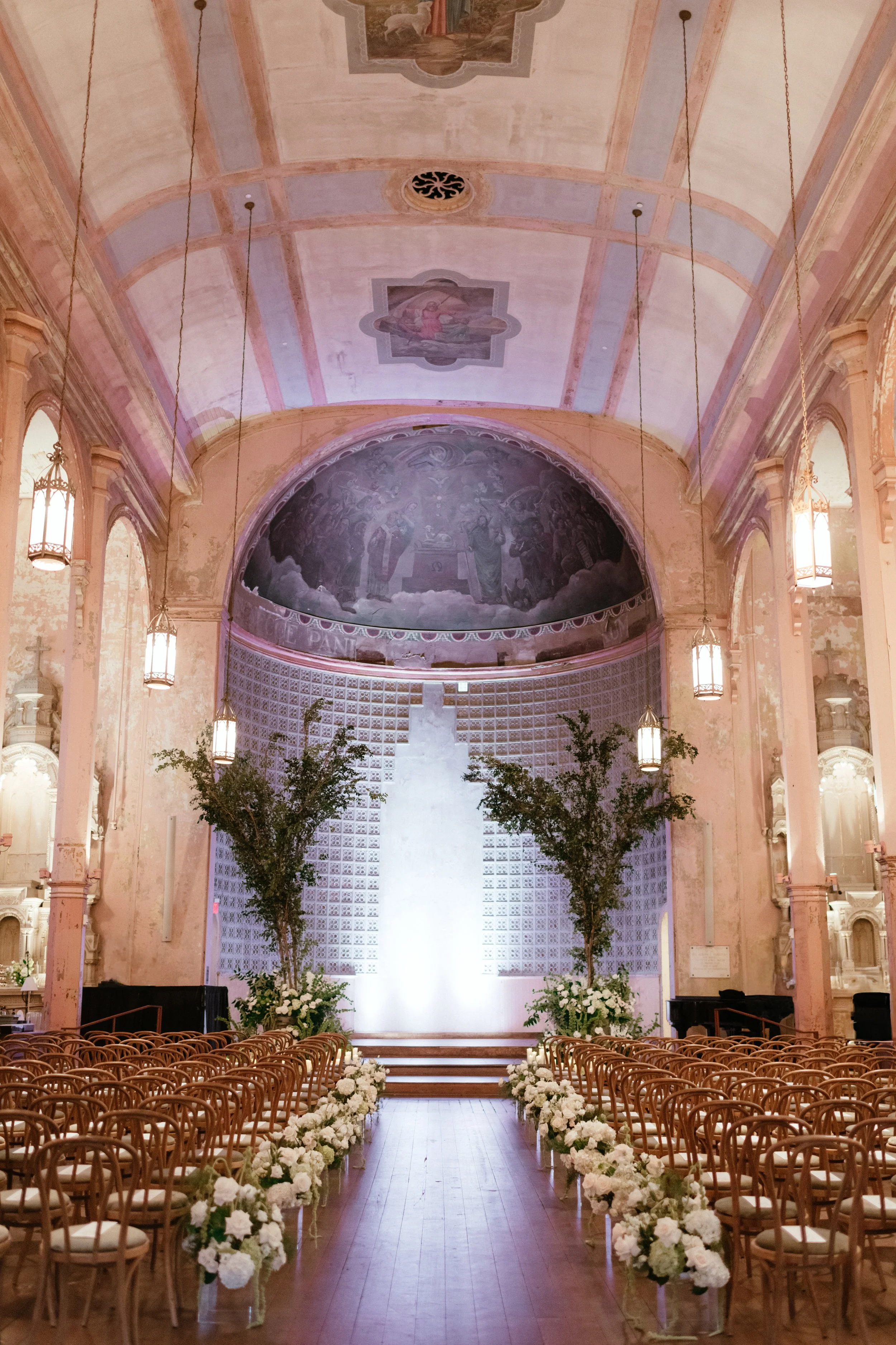 Indoor church wedding venue decorated with white flowers along the aisle and potted plants near the altar. Rows of wooden chairs are arranged on either side, leading to a lit altar with soft lighting and a large, arched wall with decorative historic 