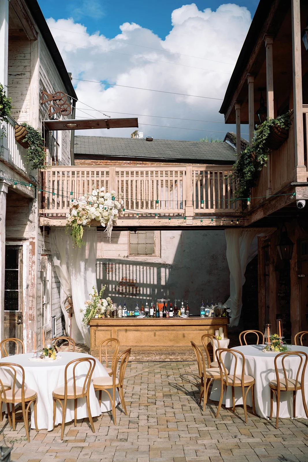 Outdoor event space with round tables covered in white tablecloths, decorated with floral centerpieces, wooden chairs, string lights, a bar with bottles, and floral arrangements under a partly cloudy sky.