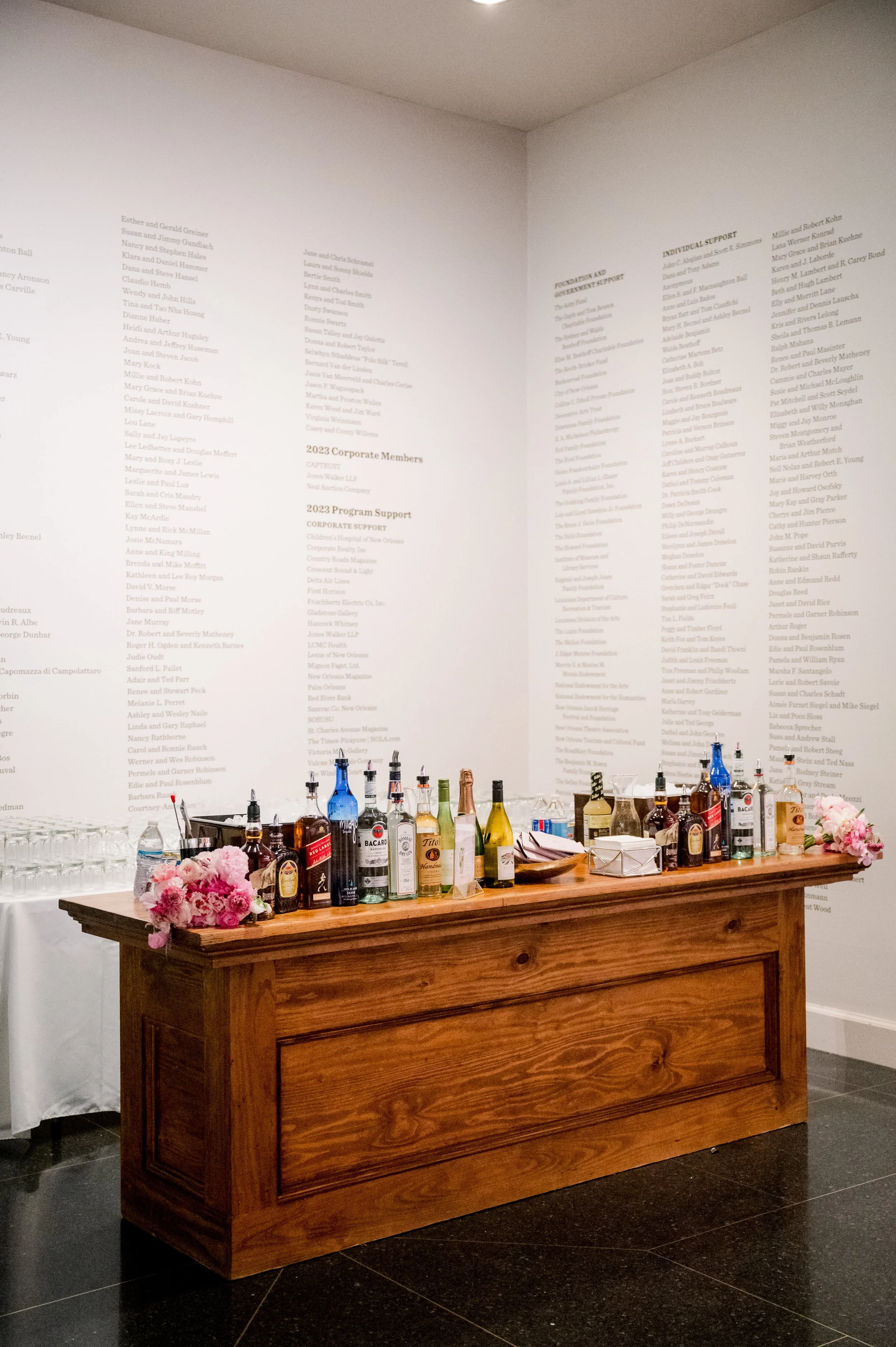 A wooden bar table with various liquor bottles, pink flowers, and empty glasses, set against a white wall with printed text.