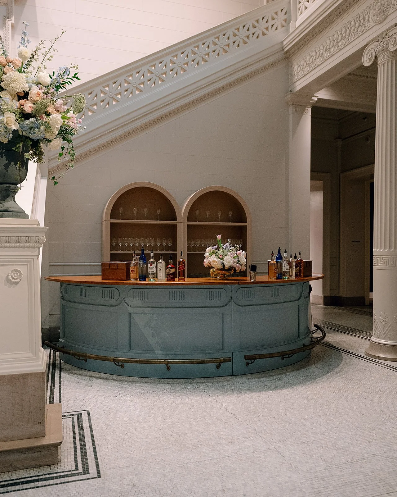 A vintage-style bar with a blue base, wooden top, and a floral centerpiece, set against a wall with two arched shelves filled with glassware, in a historic or elegant setting.