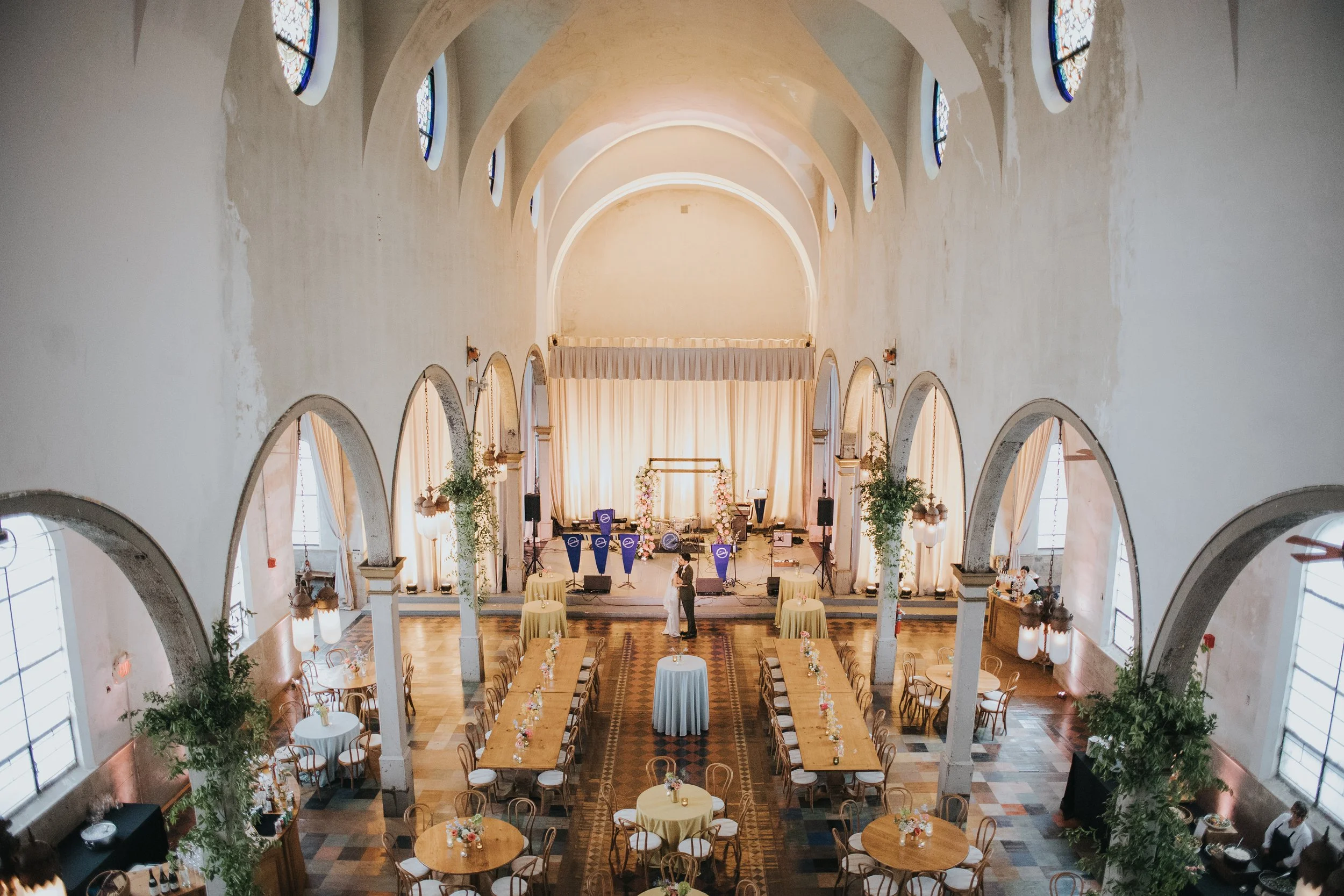 Interior of a large church or hall decorated for a wedding or event, with round tables and chairs, a stage with floral arrangements, and stained glass windows along the sides.