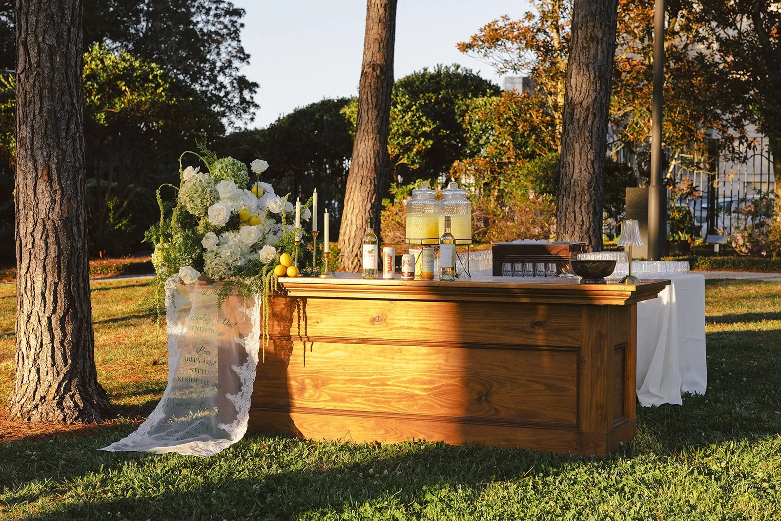 Outdoor beverage station with a wooden table, large floral arrangement, candles, bottles, and serving items, set among trees in a park or garden during late afternoon or early evening.