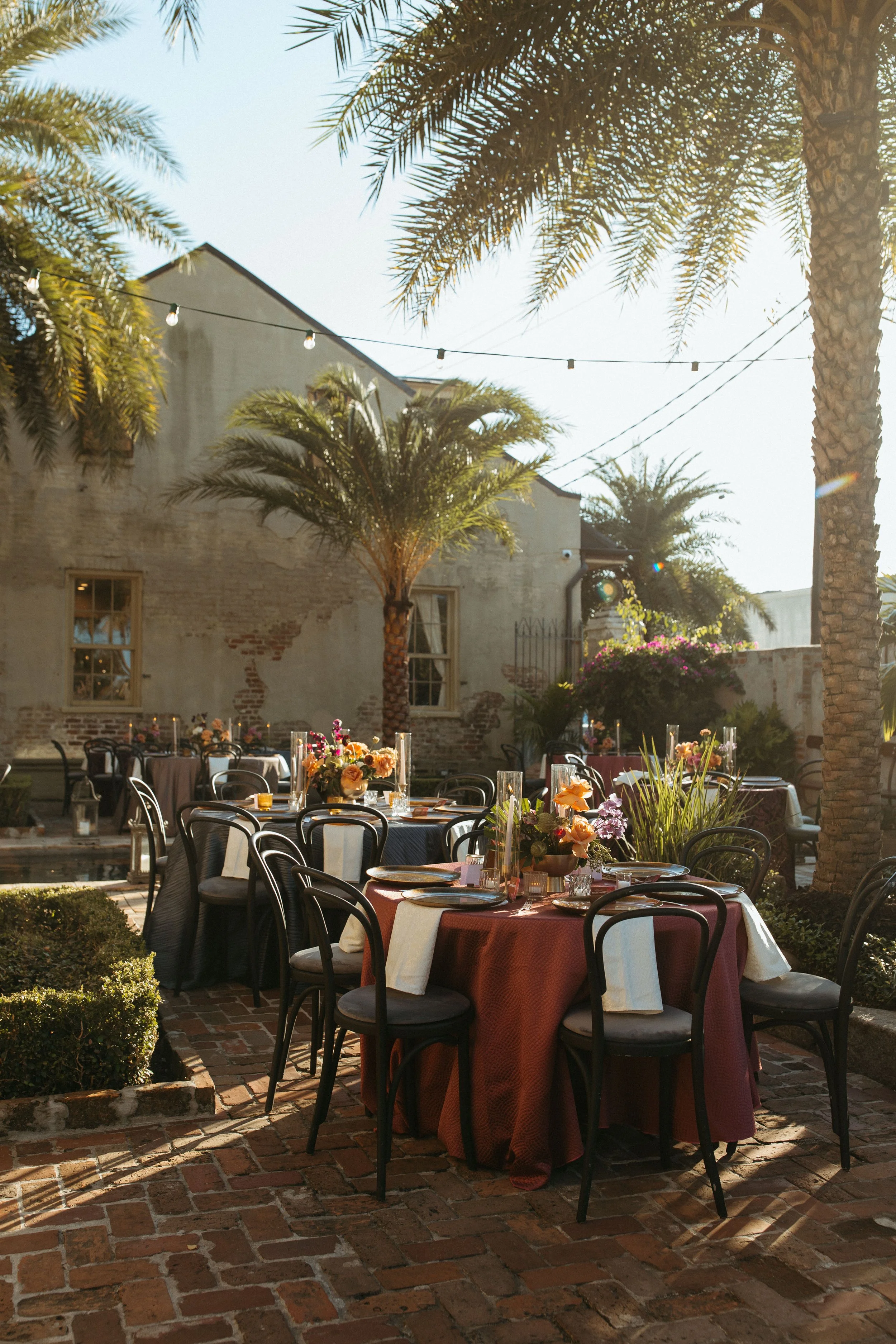 Outdoor event setup with round tables covered in burgundy and white tablecloths, decorated with floral centerpieces, surrounded by black chairs, set on a brick patio, with palm trees, string lights, and a weathered brick wall in the background.