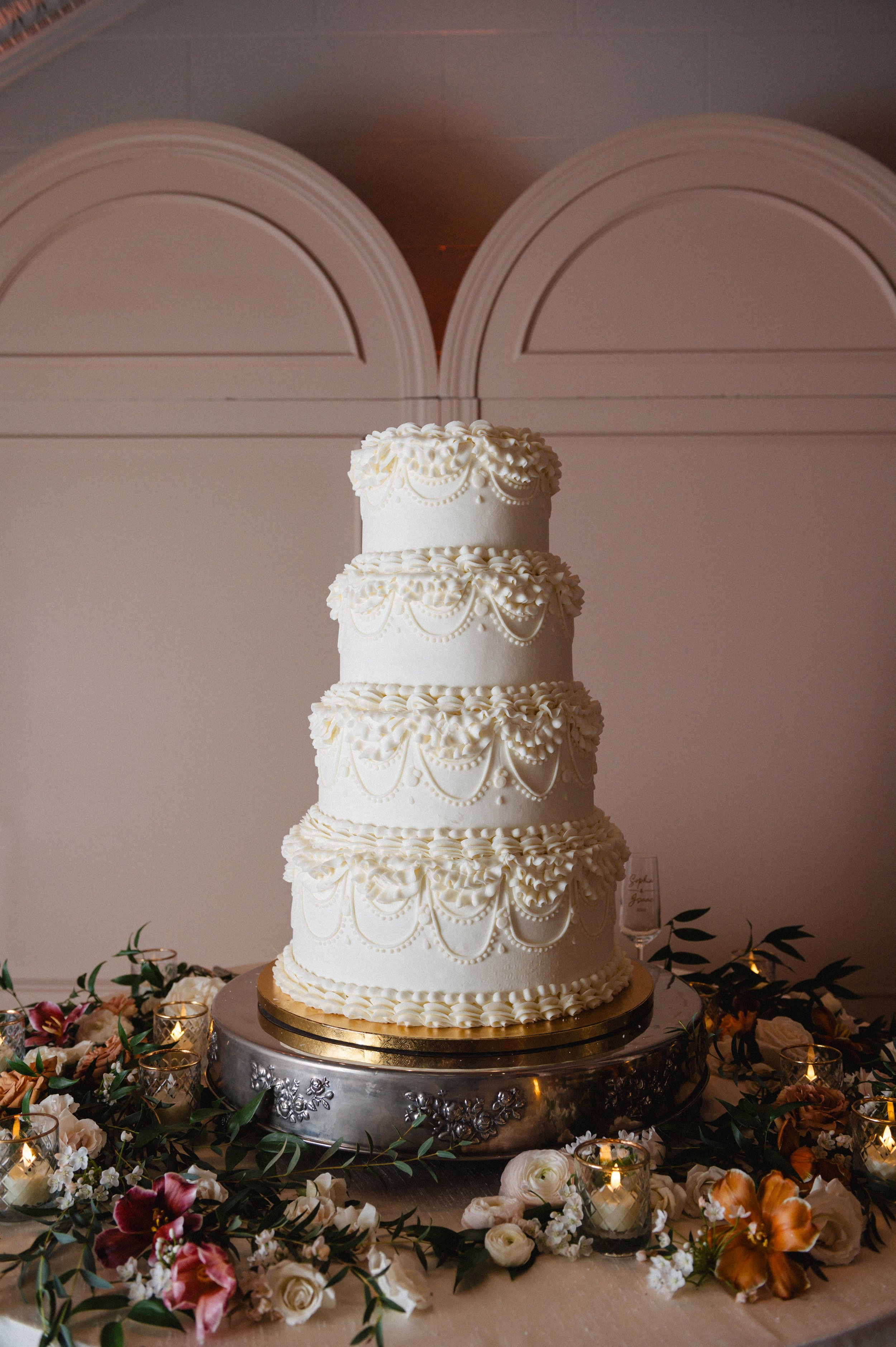 A tall, ornate white wedding cake with multiple tiers decorated with piped icing patterns. The cake is placed on a silver stand surrounded by flowers and small lit candles on a table.