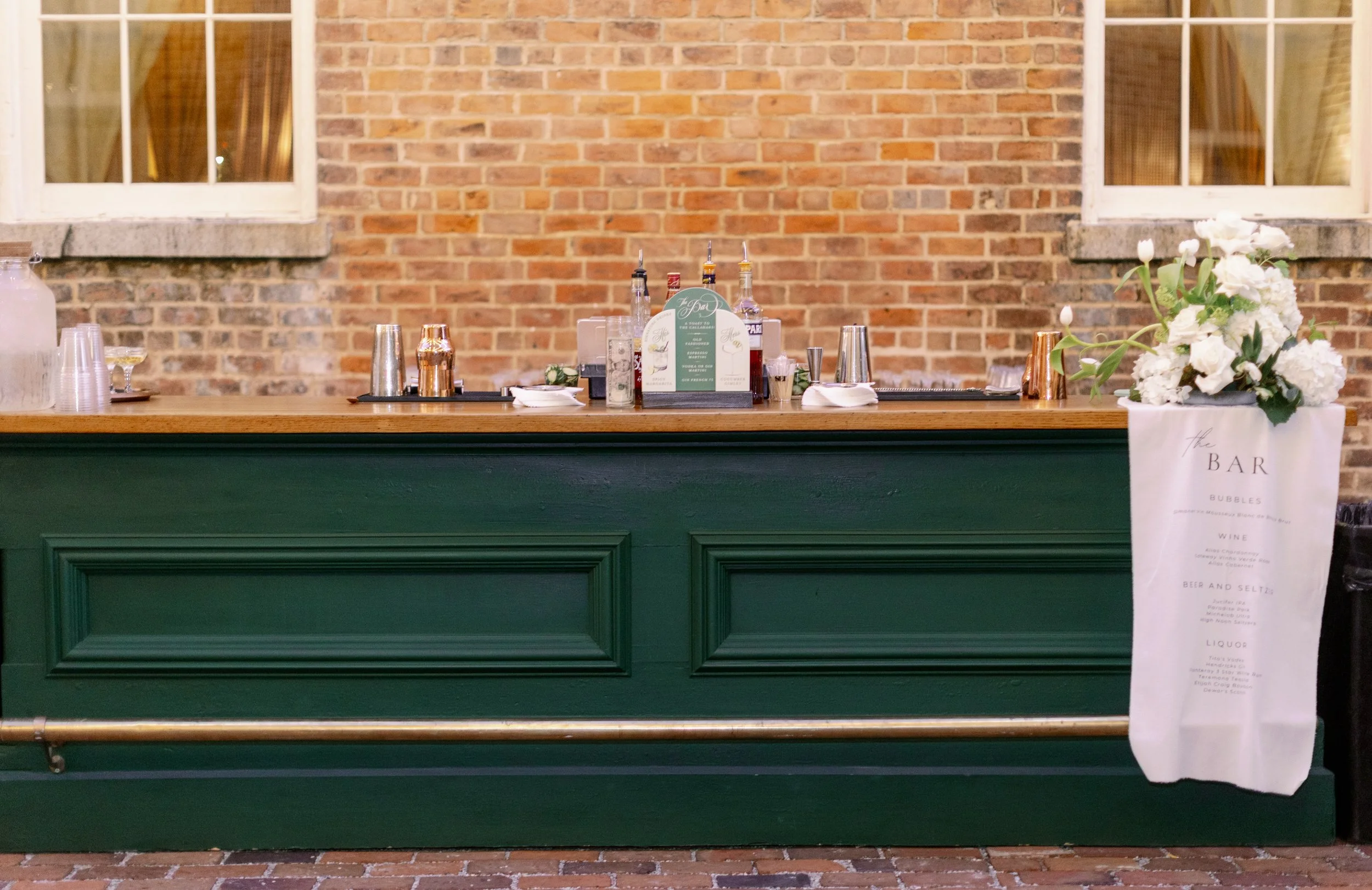 Bar area with a brick wall background, a wooden bar counter painted dark green, and a flower arrangement on a white paper bag with menu items displayed behind the bar.