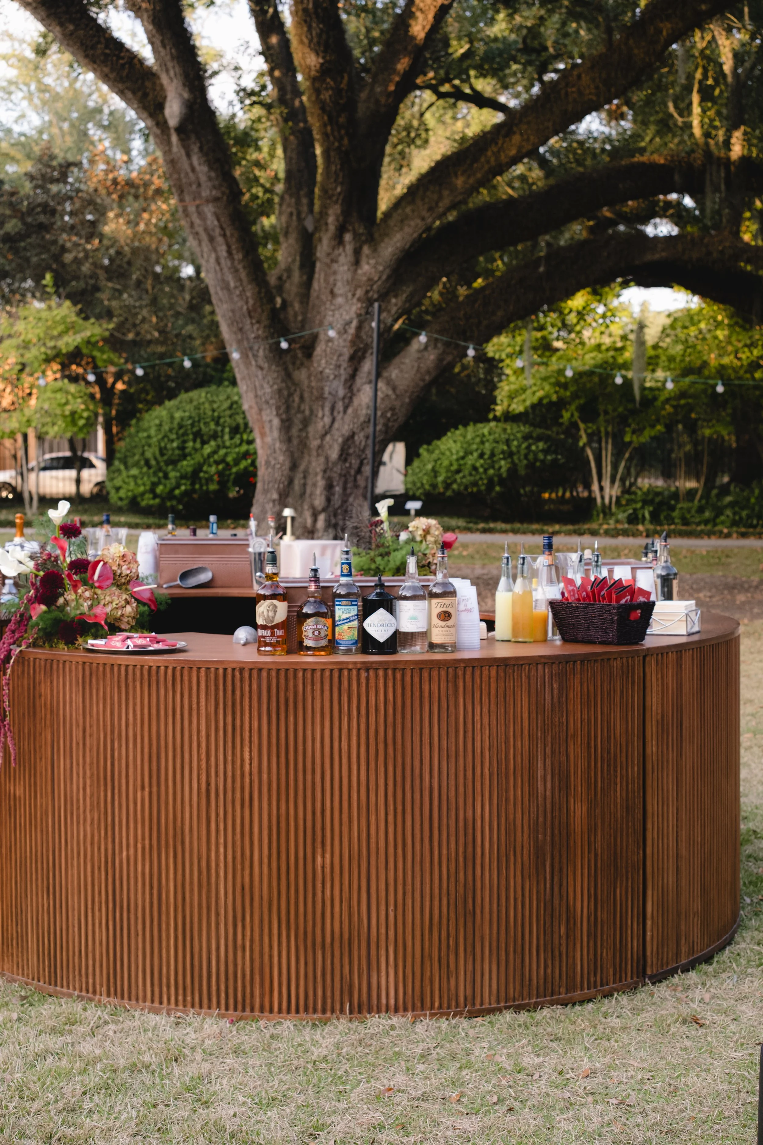 A round wooden bar table set up outdoors in a yard with trees and bushes, decorated with bottles of liquor, cocktail mixers, a flower bouquet, and a basket of red napkins, with a large tree in the background.