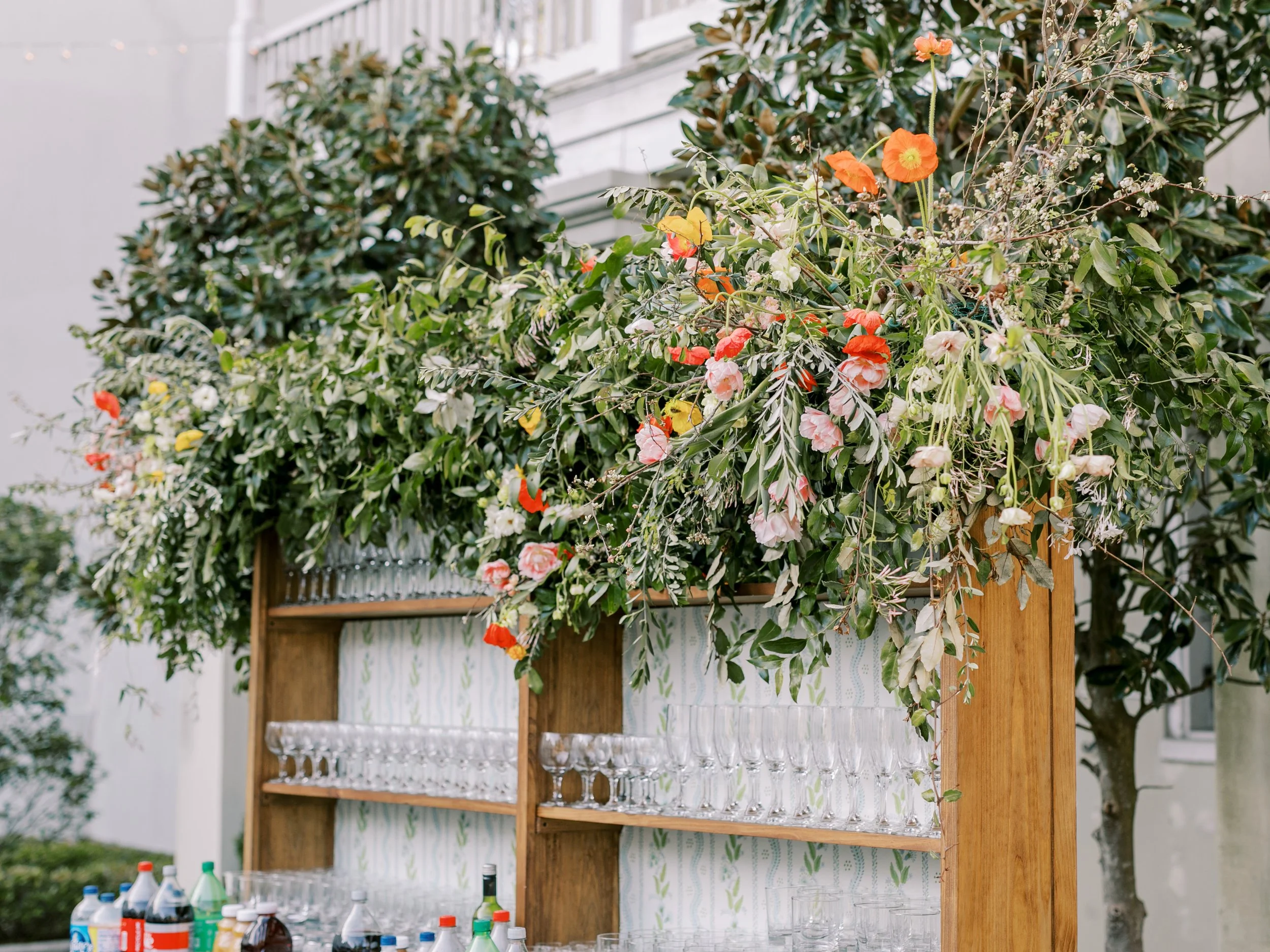 An outdoor bar setup with wooden shelves holding glasses, bottles, and drinks, decorated with lush green foliage and vibrant orange, pink, and white flowers.
