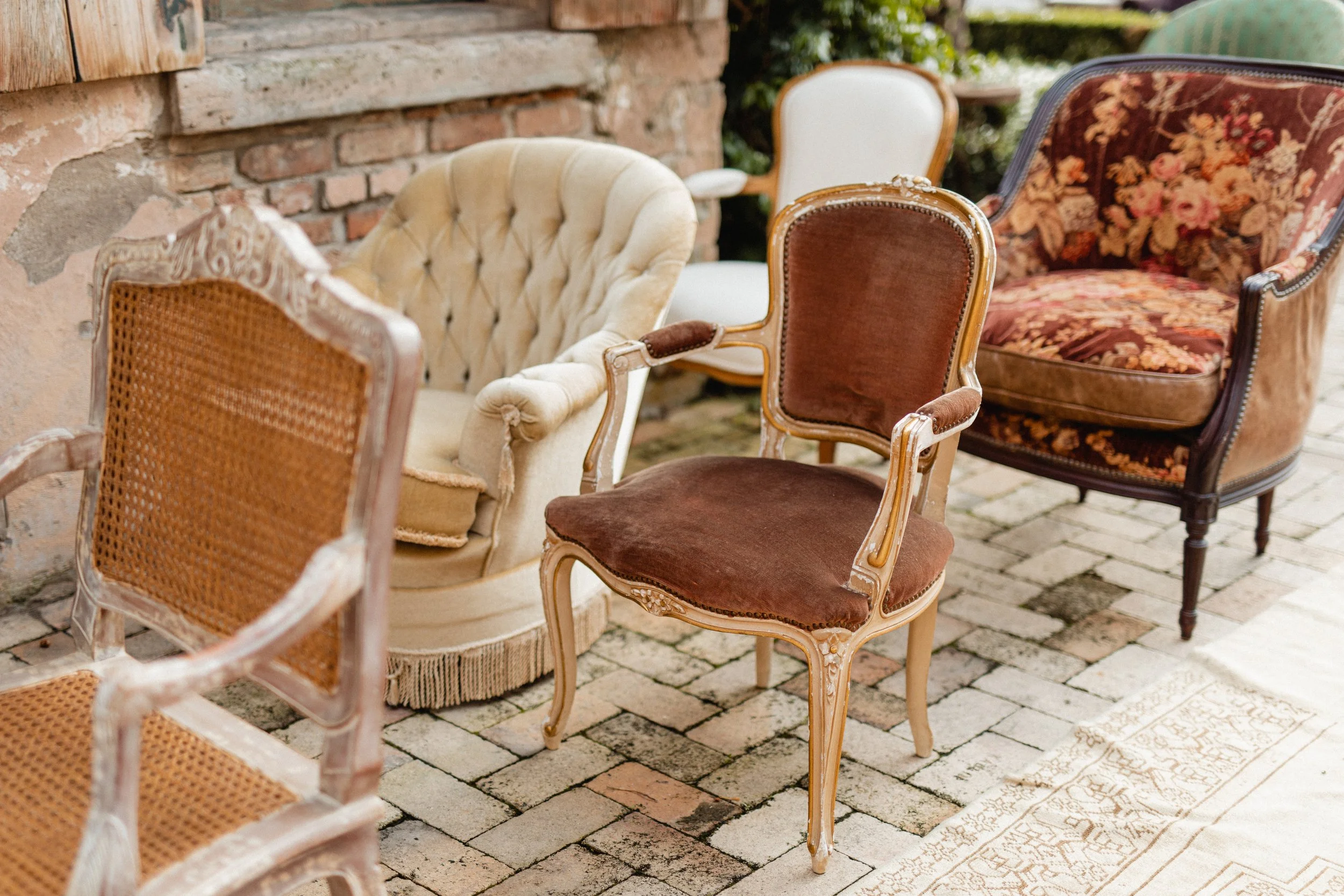 Various vintage and antique chairs placed outdoors on a stone patio near a brick wall and greenery.