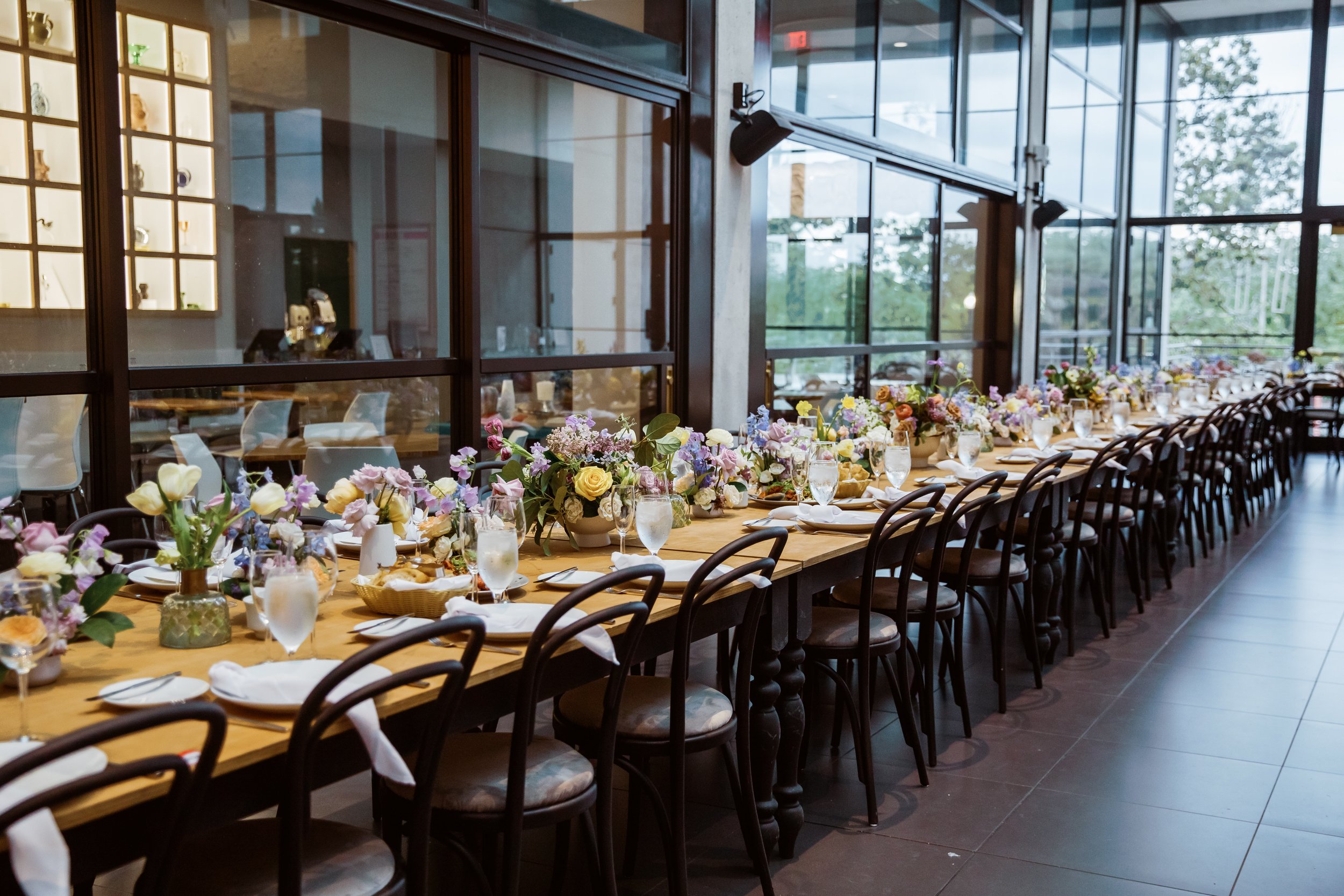 A large dining table set for a formal event with floral centerpieces, glass water goblets, white napkins, and bread rolls, inside a modern building with large glass windows and a view of trees outside.